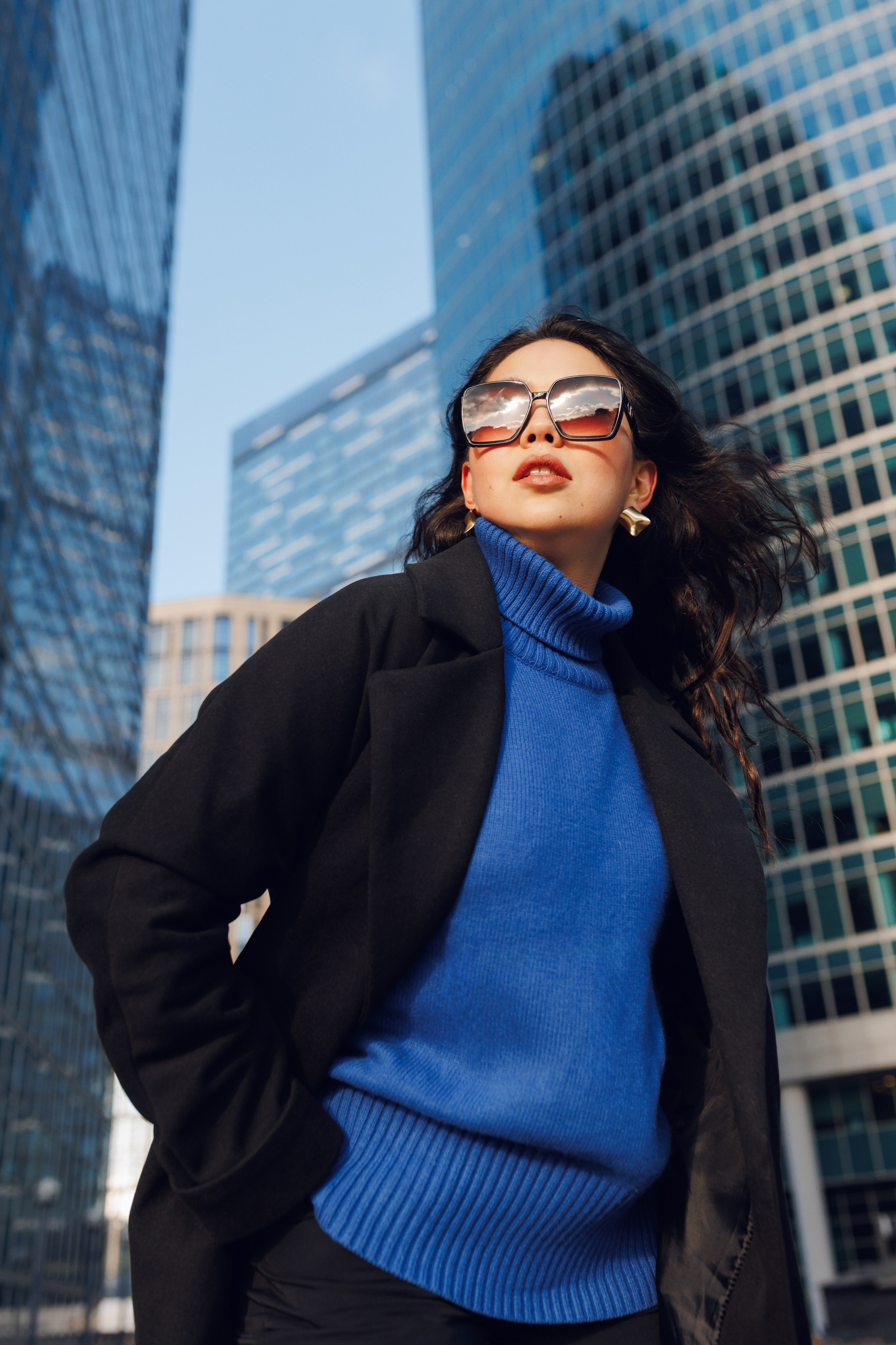 Девушка в очках и пальто позирует на фоне делового центра Москва-Сити.