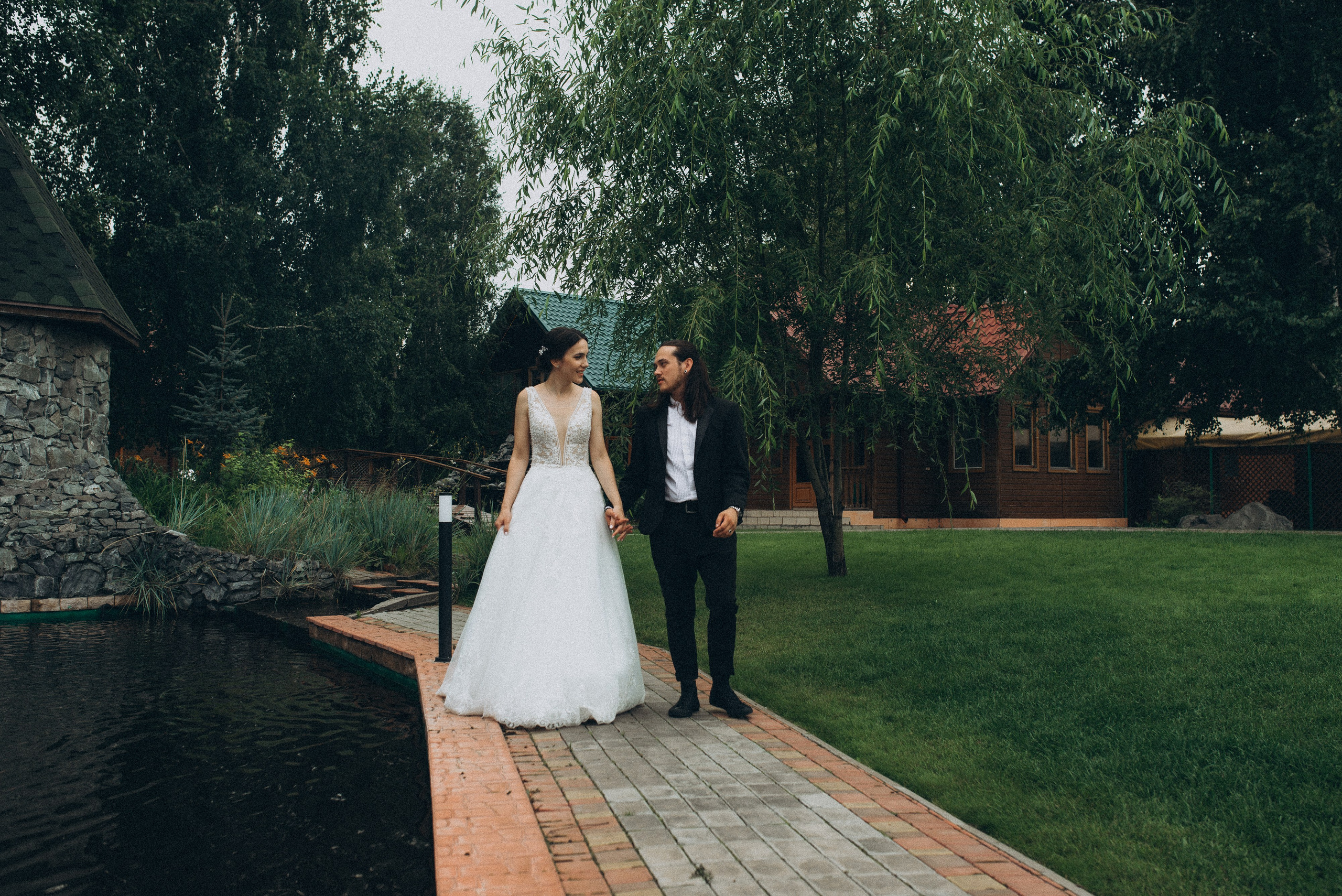 Жених Никита и невеста Линда идут по дорожке, держась за руки, на фоне деревянных домиков. Свадебная прогулка за городом.