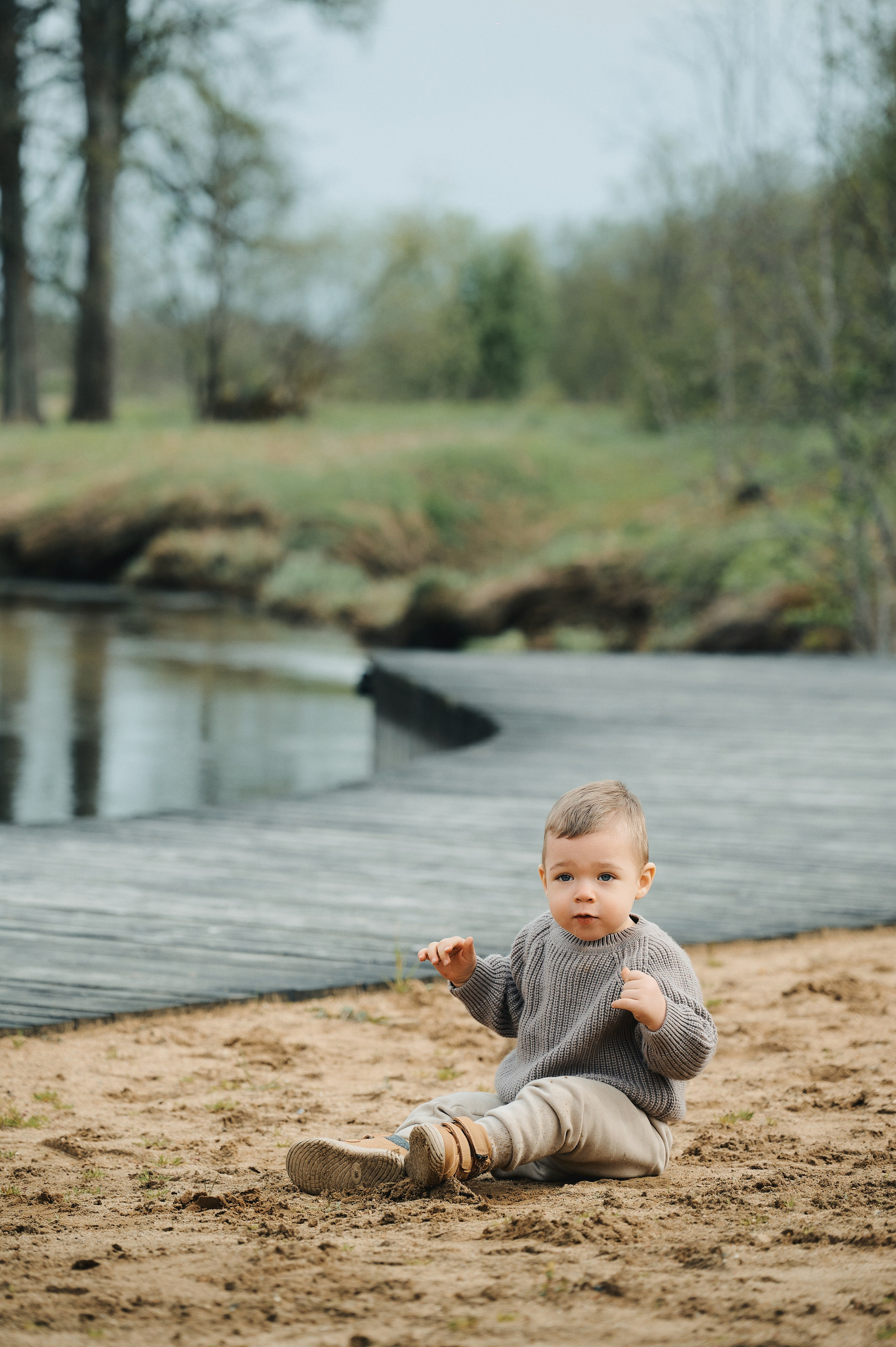 мальчик сидит на песке у реки, вязаный свитер и светлые брюки, фон — деревянная набережн