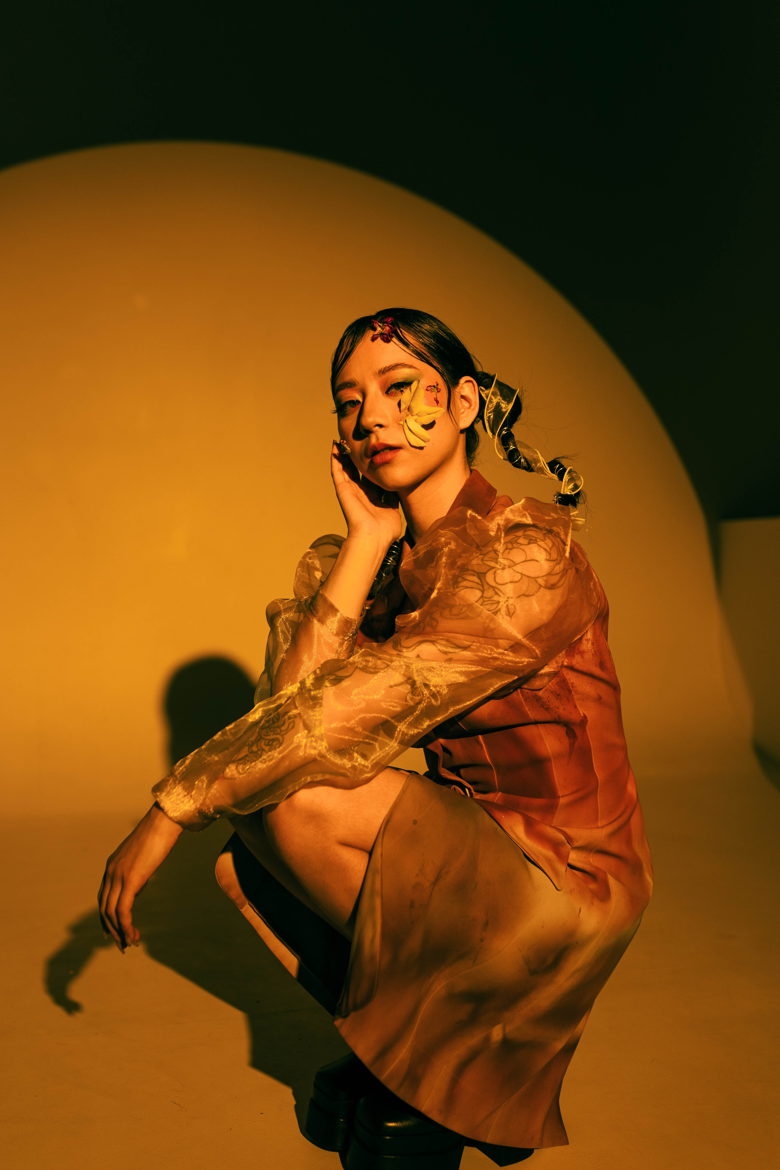 Молодая женщина с ярким цветочным макияжем на лице сидит на корточках в эффектной позе. Теплый желтый фон подчеркивает ее художественную выразительность и модный наряд.