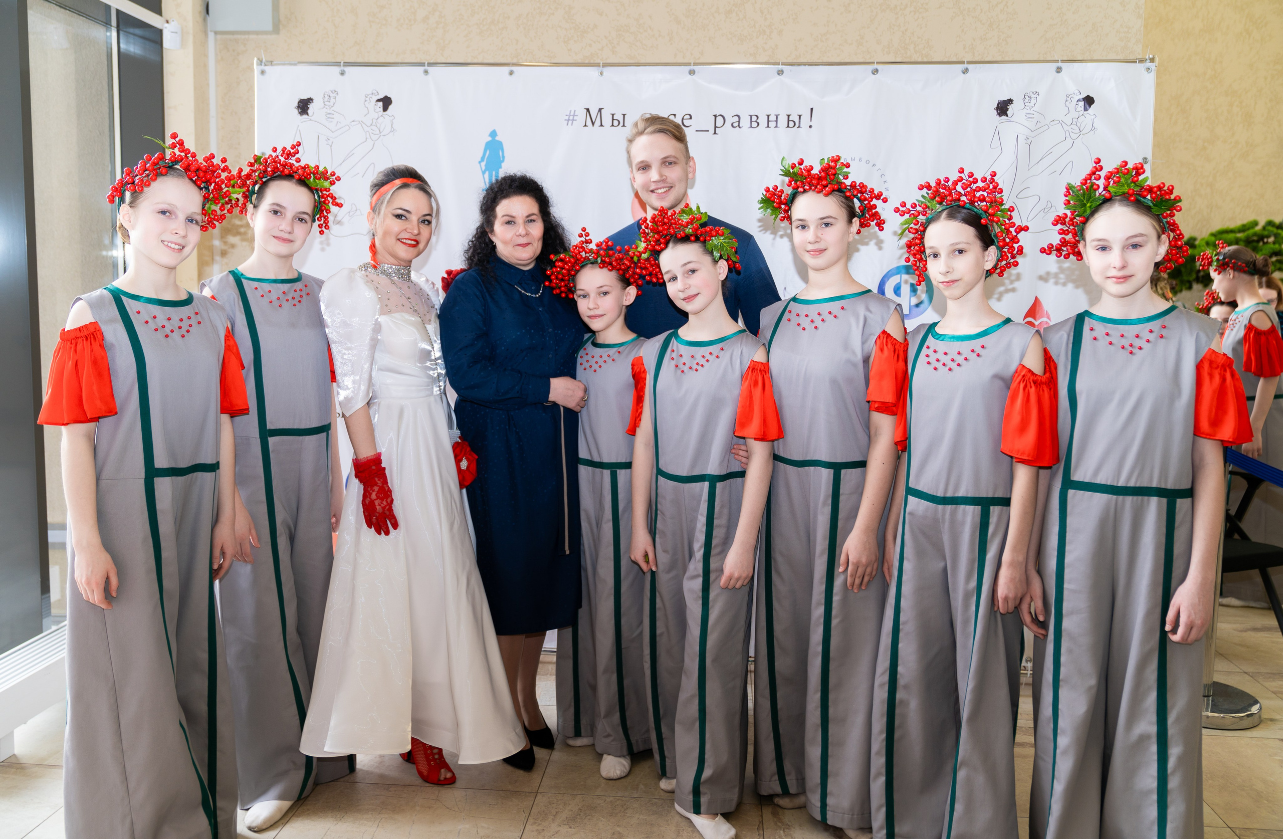 Групповое фото участников инклюзивного бала «Мы все равны». Женщина в белом платье, дети в серых костюмах с венками из красных ягод и взрослые. Фотограф Мальцев Евгений.