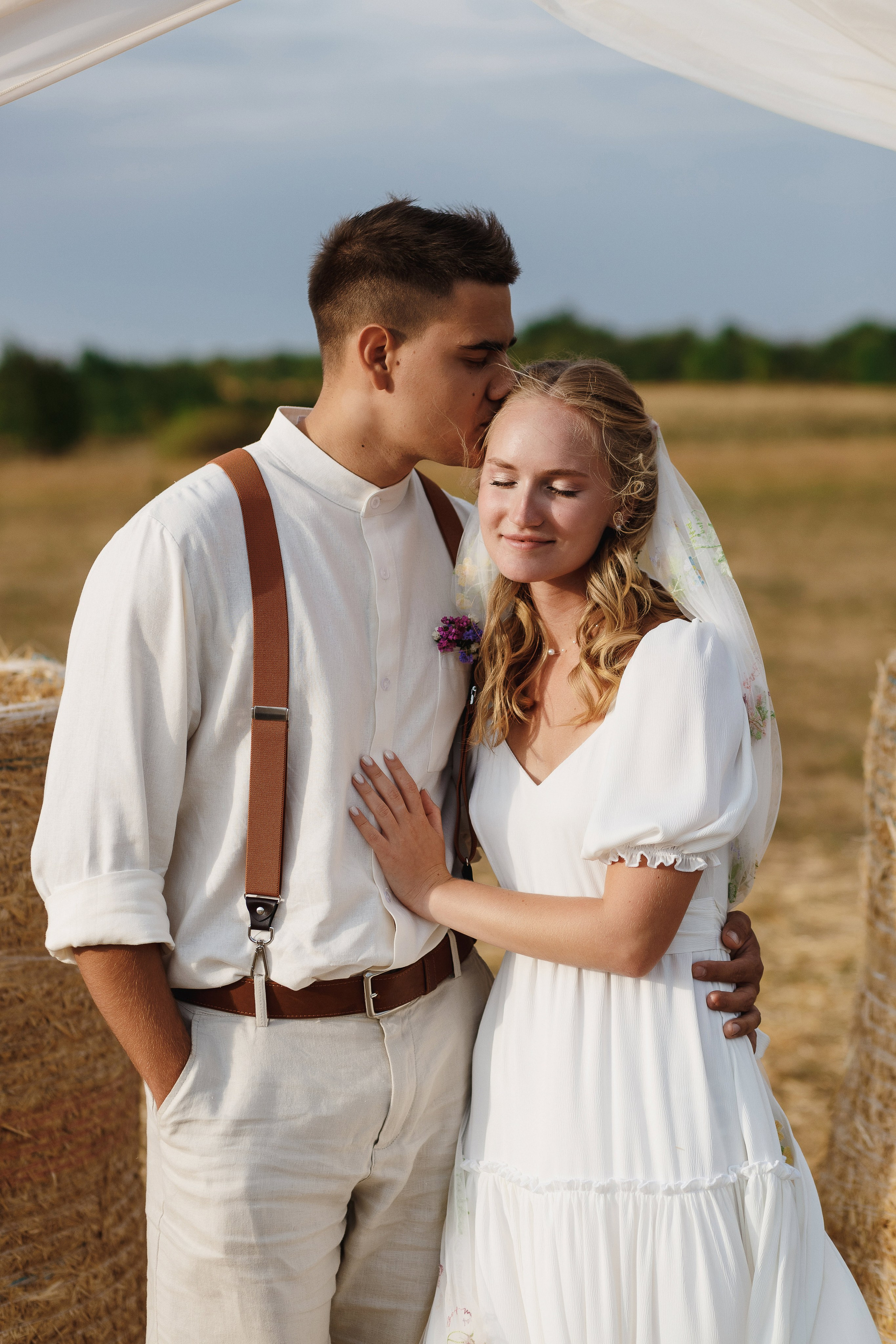 Жених целует невесту в лоб на фоне сена и свадебной арки — свадебная фотосессия, фотограф Таганрог