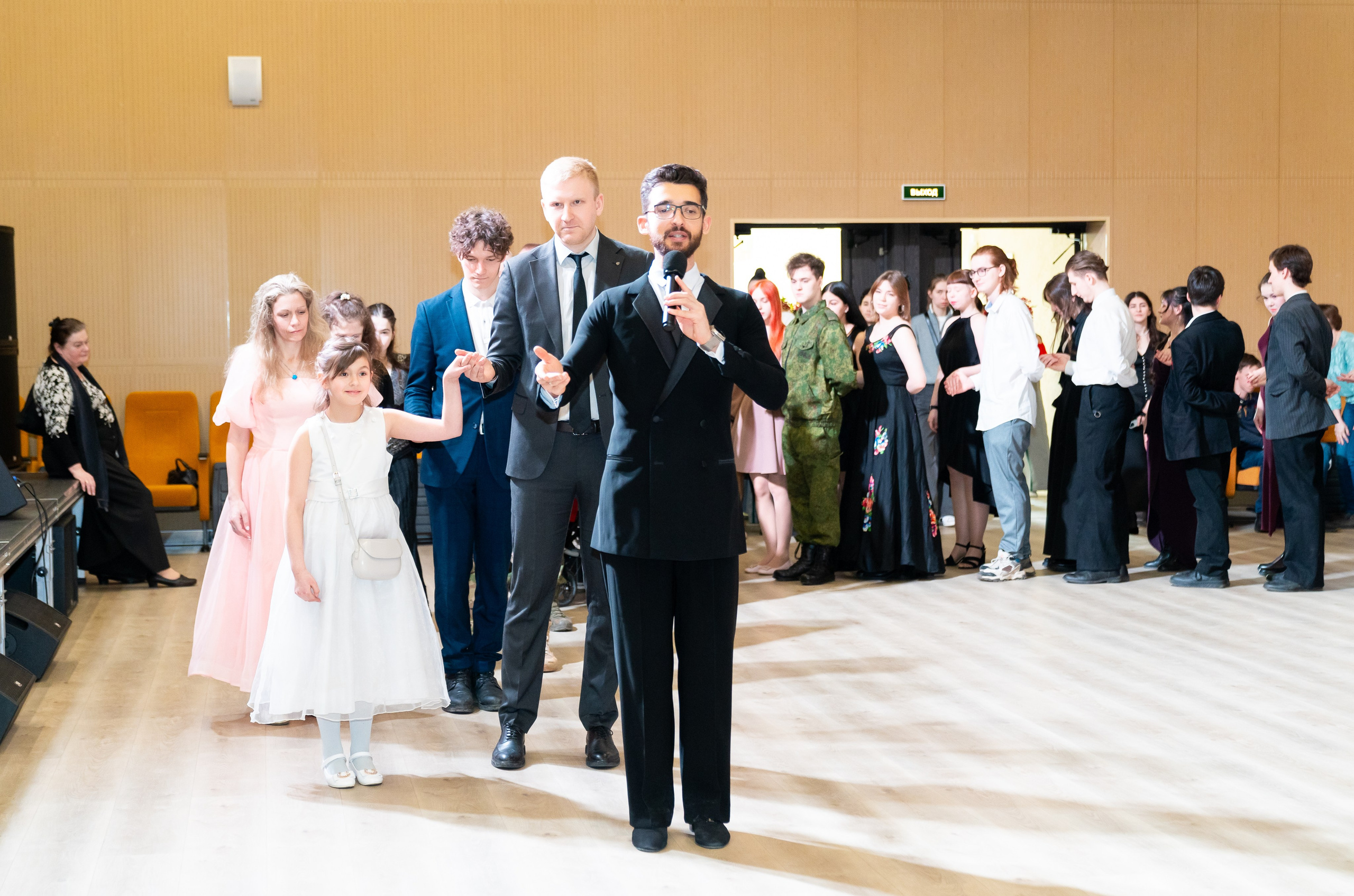 Ведущий в чёрном костюме с микрофоном открывает инклюзивный бал проекта «Мы все равны». Рядом девочка в белом платье и другие участники. Фотограф Мальцев Евгений.