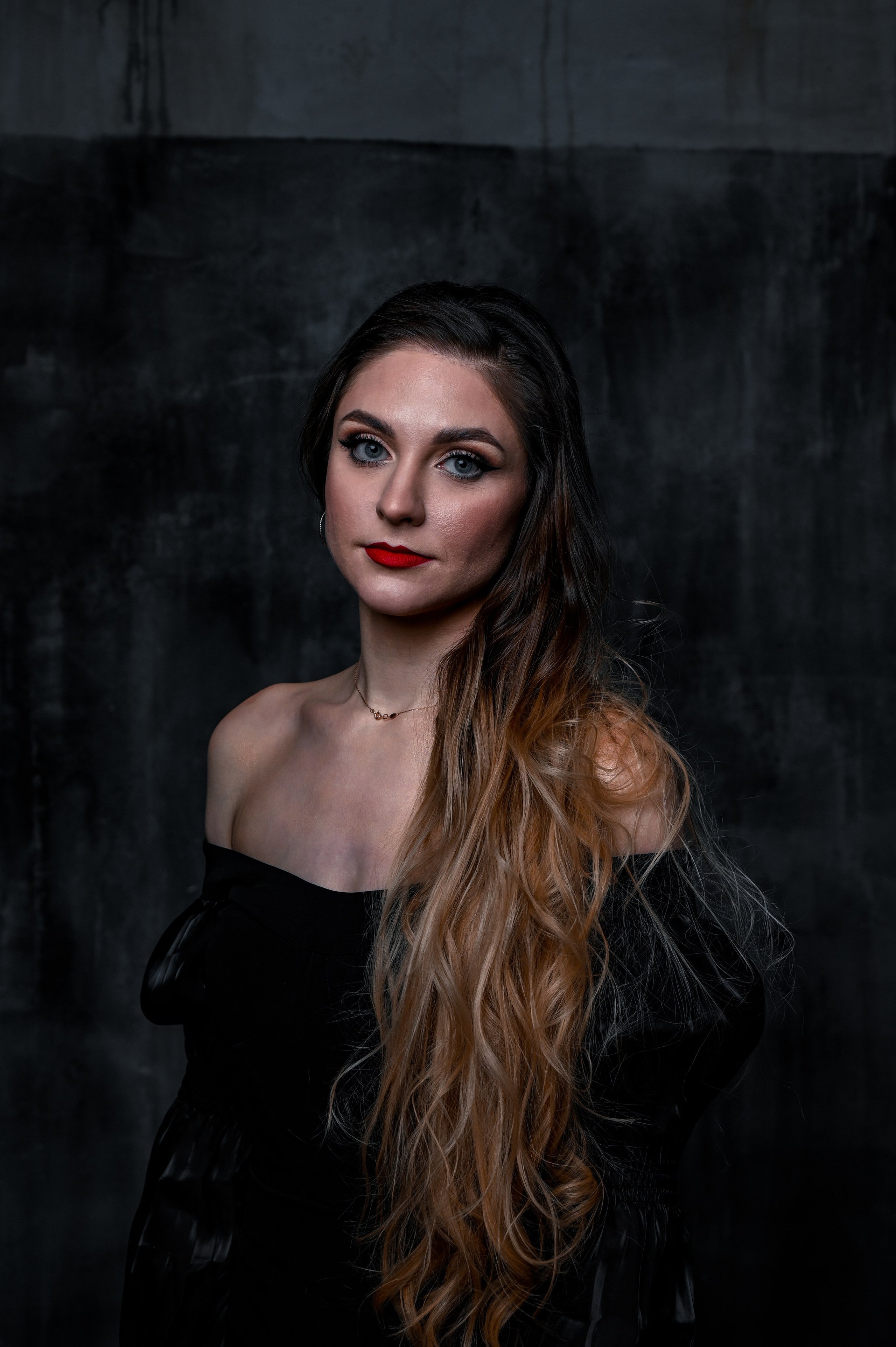 Эмоциональный женский портрет в стиле нуар с естественным освещением. Фотография подчёркивает глубину взгляда и утончённость молодой женщины с длинными волнистыми волосами, украшенными мягкими светлыми прядями. Модель в чёрном платье с открытыми плечами и изящным ожерельем создаёт элегантный и загадочный образ. Идеально для личного блога, соцсетей или рекламных проектов. Профессиональная съёмка в студии с акцентом на естественную красоту.