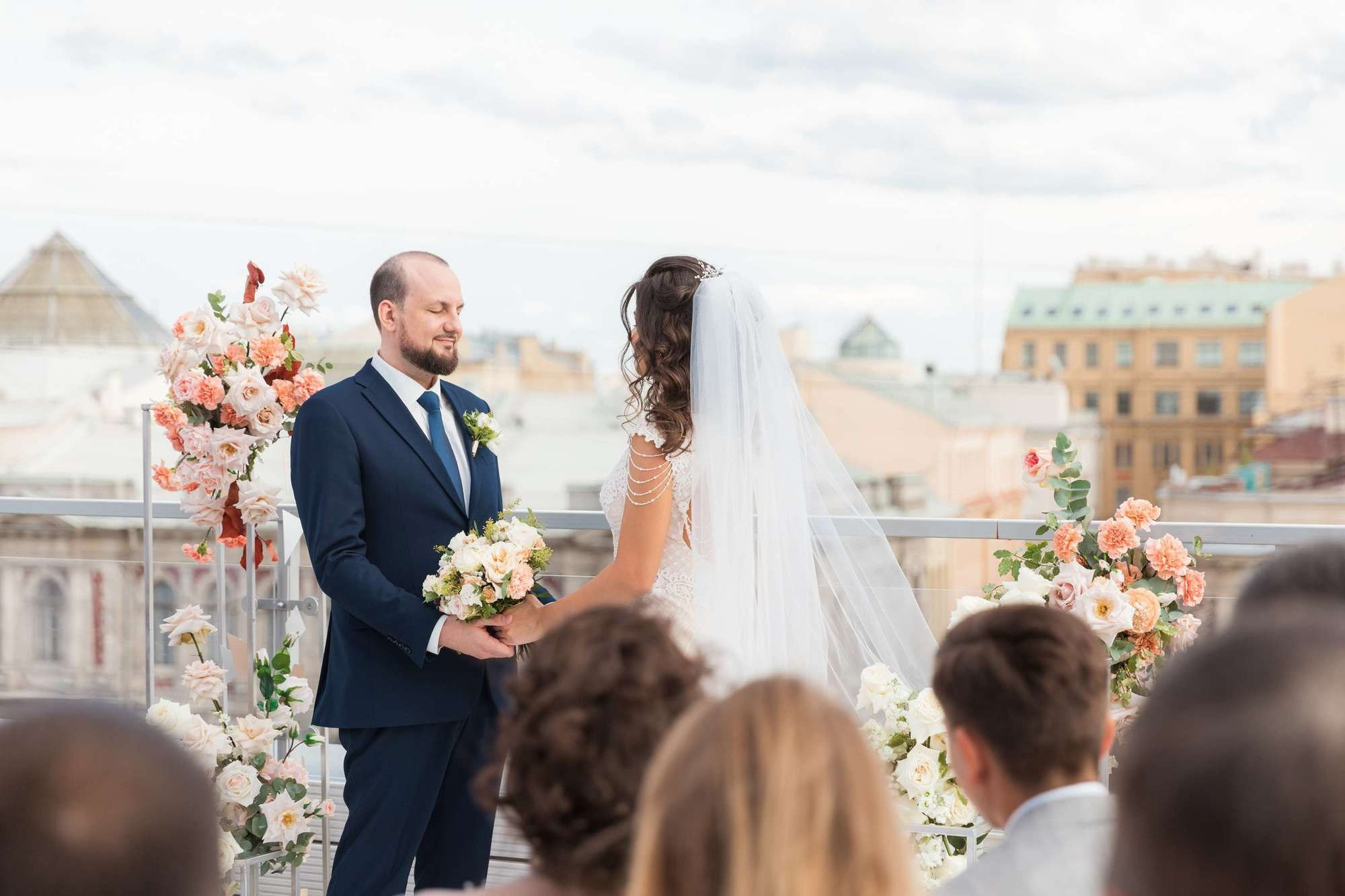 Молодожены держатся за руки у арки из цветов на крыше в СПб