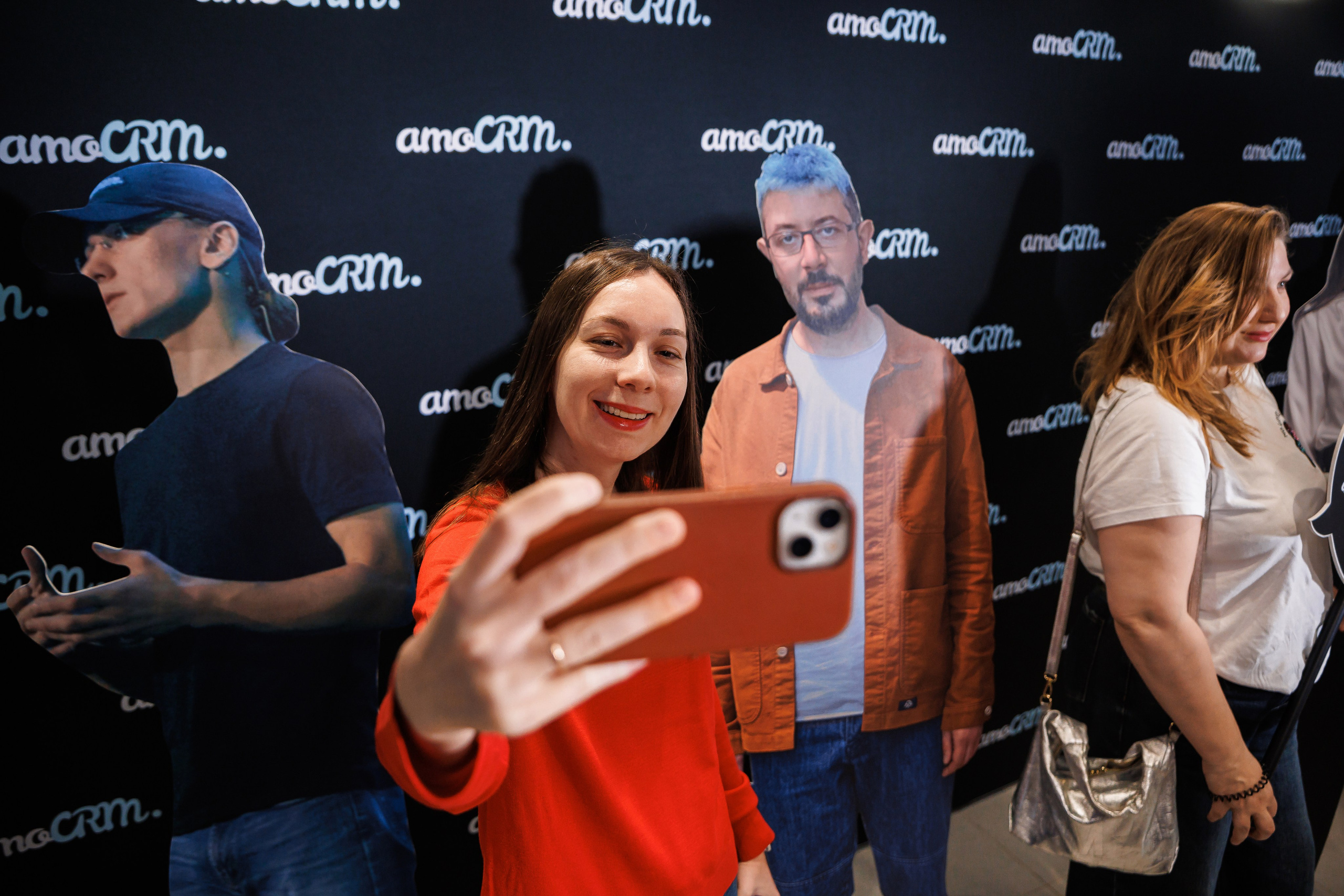 Женщина в красной рубашке делает селфи на фоне картонных фигур людей в натуральную величину на мероприятии, где на заднем плане повторяется логотип amoCRM. Рядом видны другие участники.