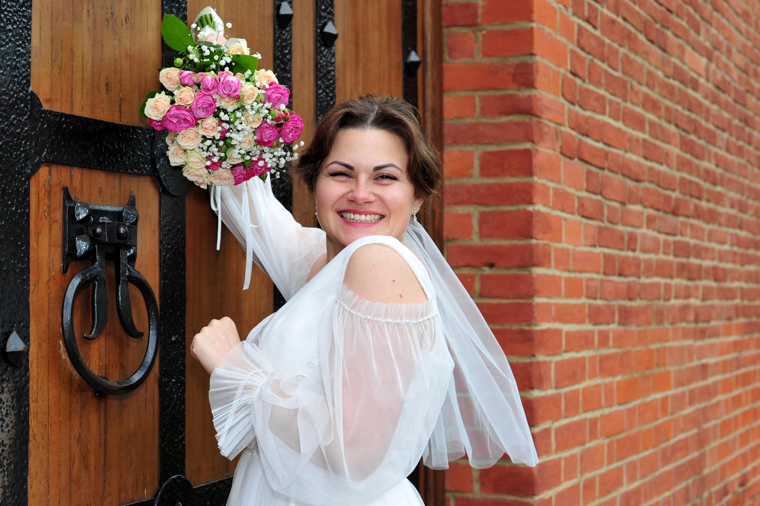 Свадьба в Царицыно пасмурным летним днем, портрет невесты со свадебным букетом на фоне старинной деревянной двери и стены из красного кирпича 