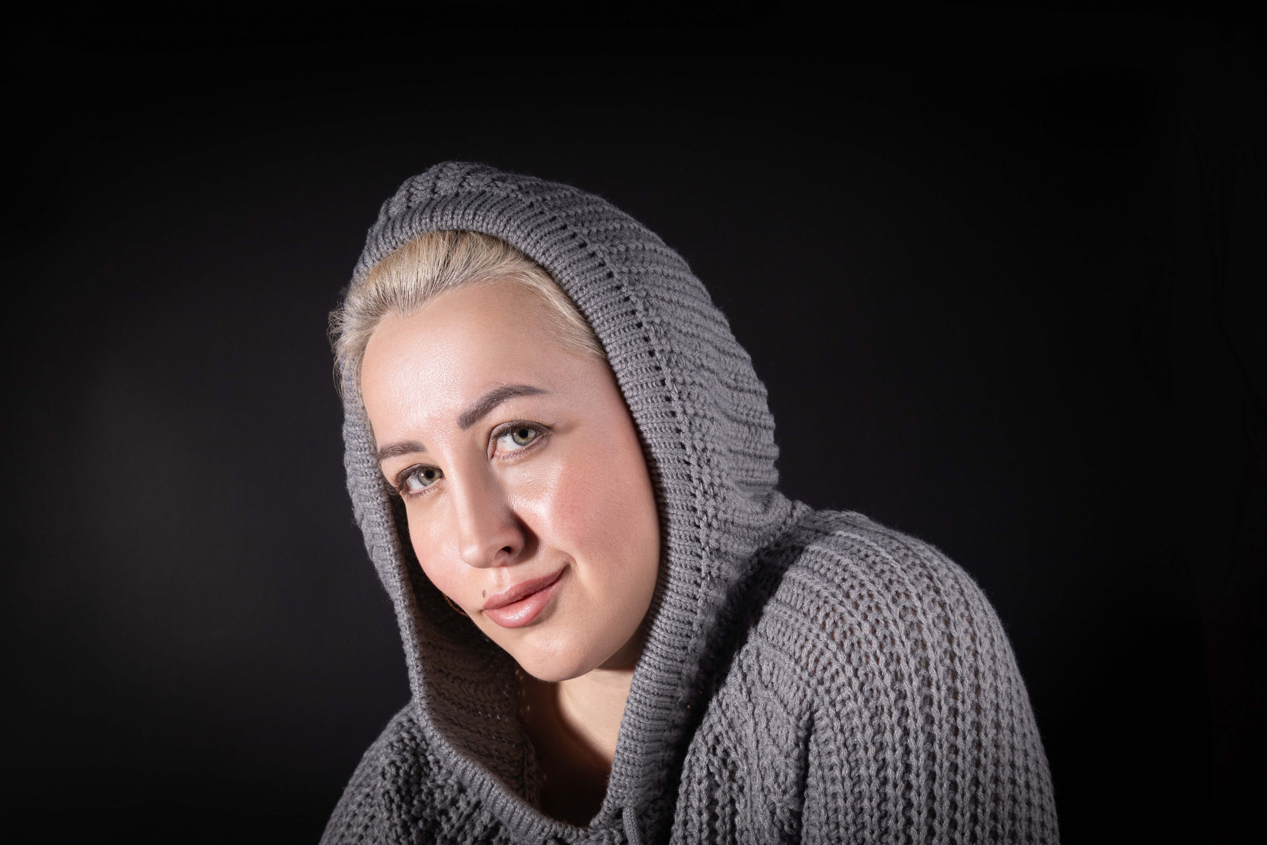 Девушка в сером вязаном свитере с капюшоном, тёмный фон.