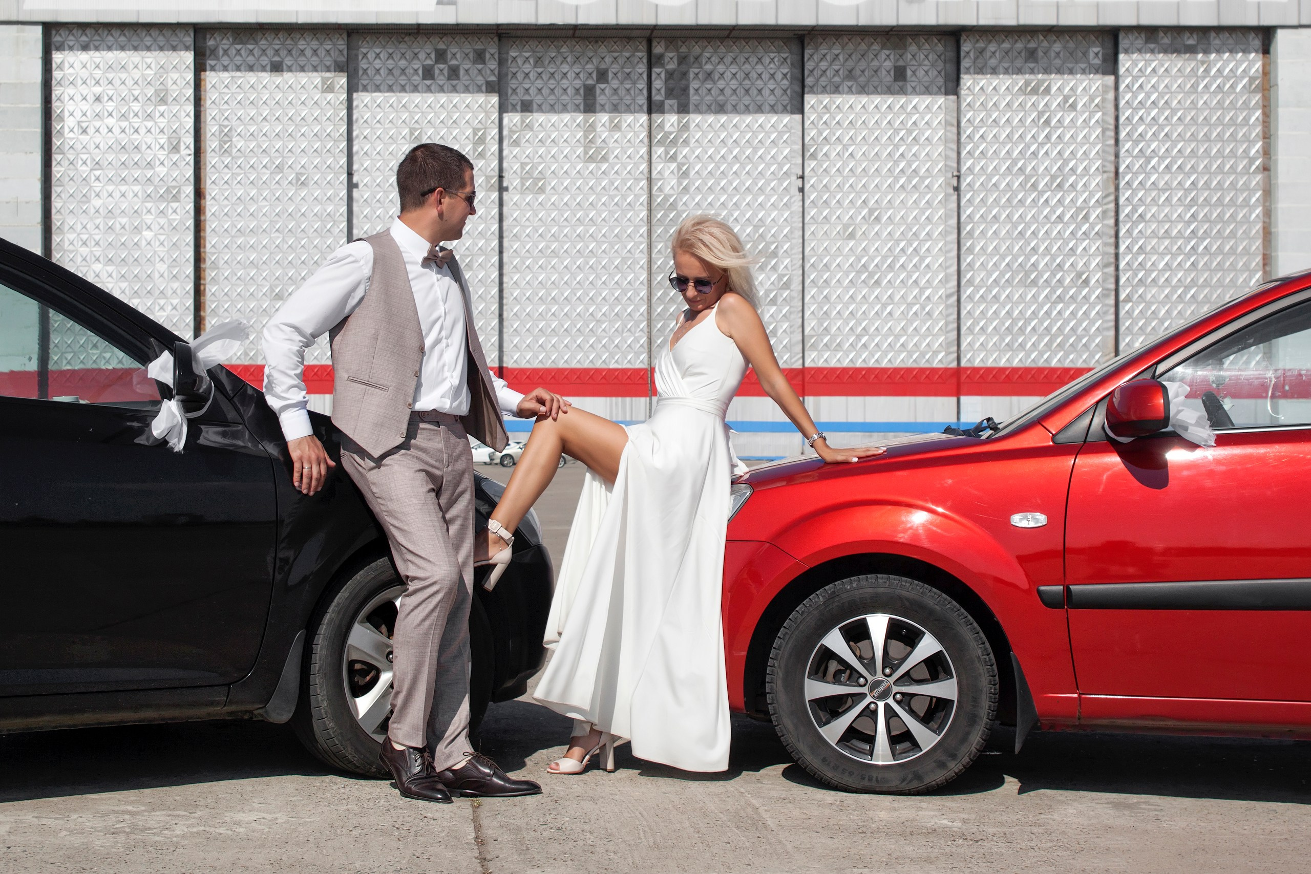 Жених и невеста около машин красного и черного цвета, невеста опирается ногой на колесо черной машины.