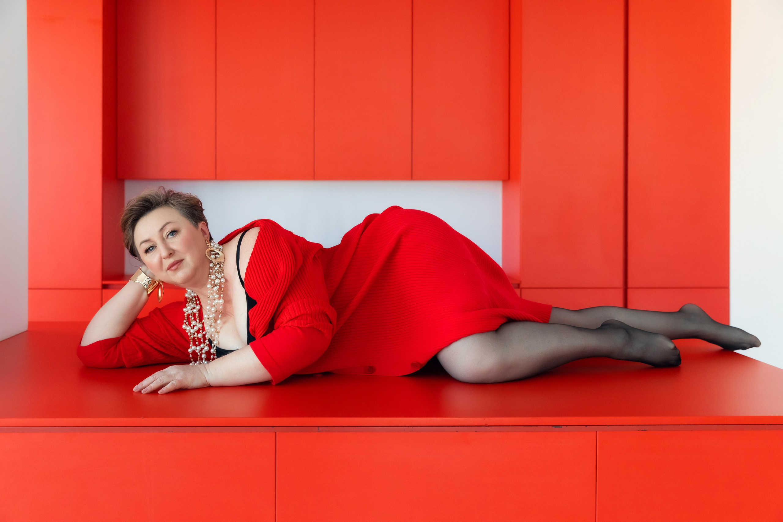 Женщина в кружевном черном платье, позирует на красном диване, акцент на украшениях и взгляде в камеру.