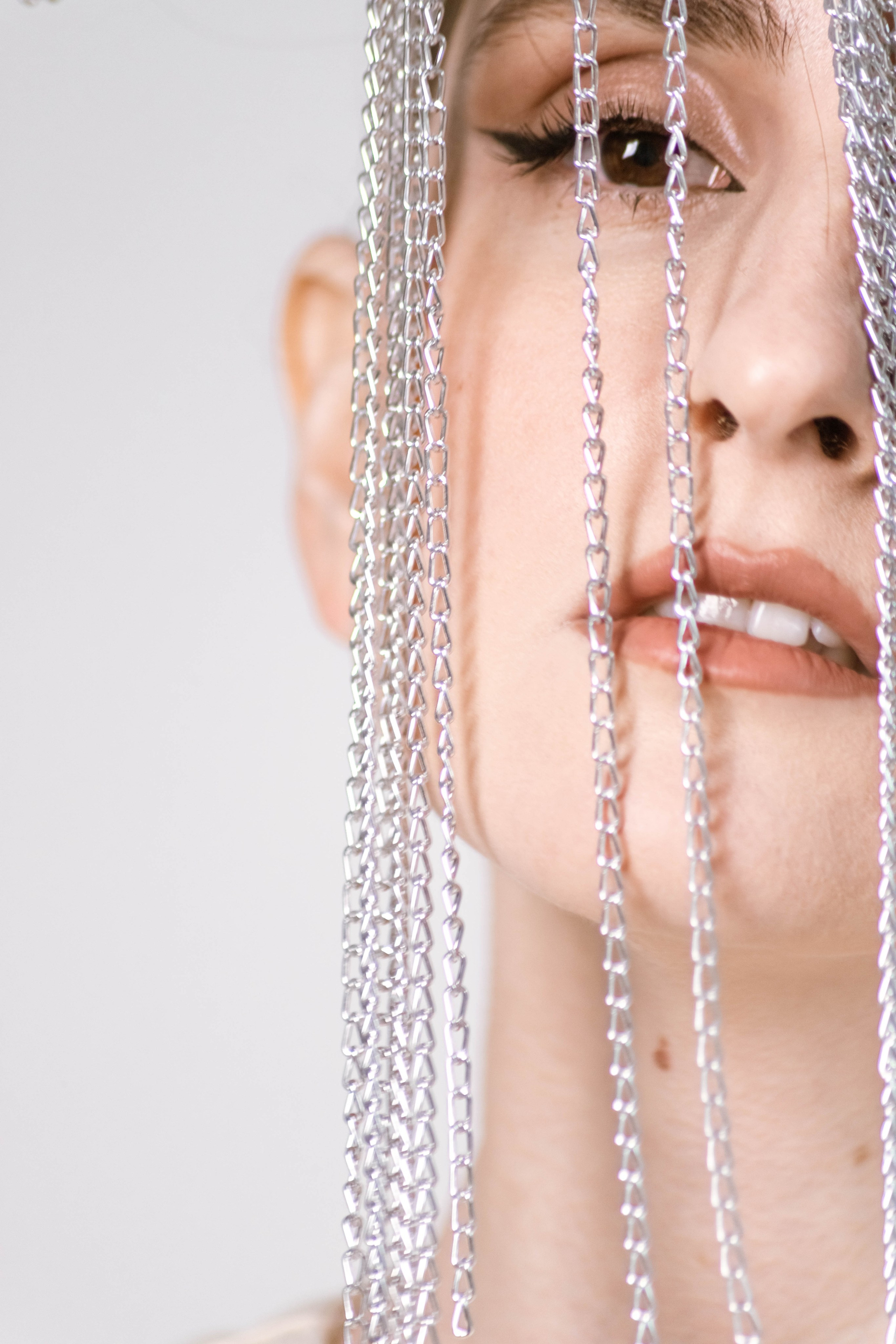 Женщина демонстрирует уникальный артистичный образ с серебряными цепочками, ниспадающими каскадом на ее лицо, подчеркивая ее поразительные черты на светлом фоне.