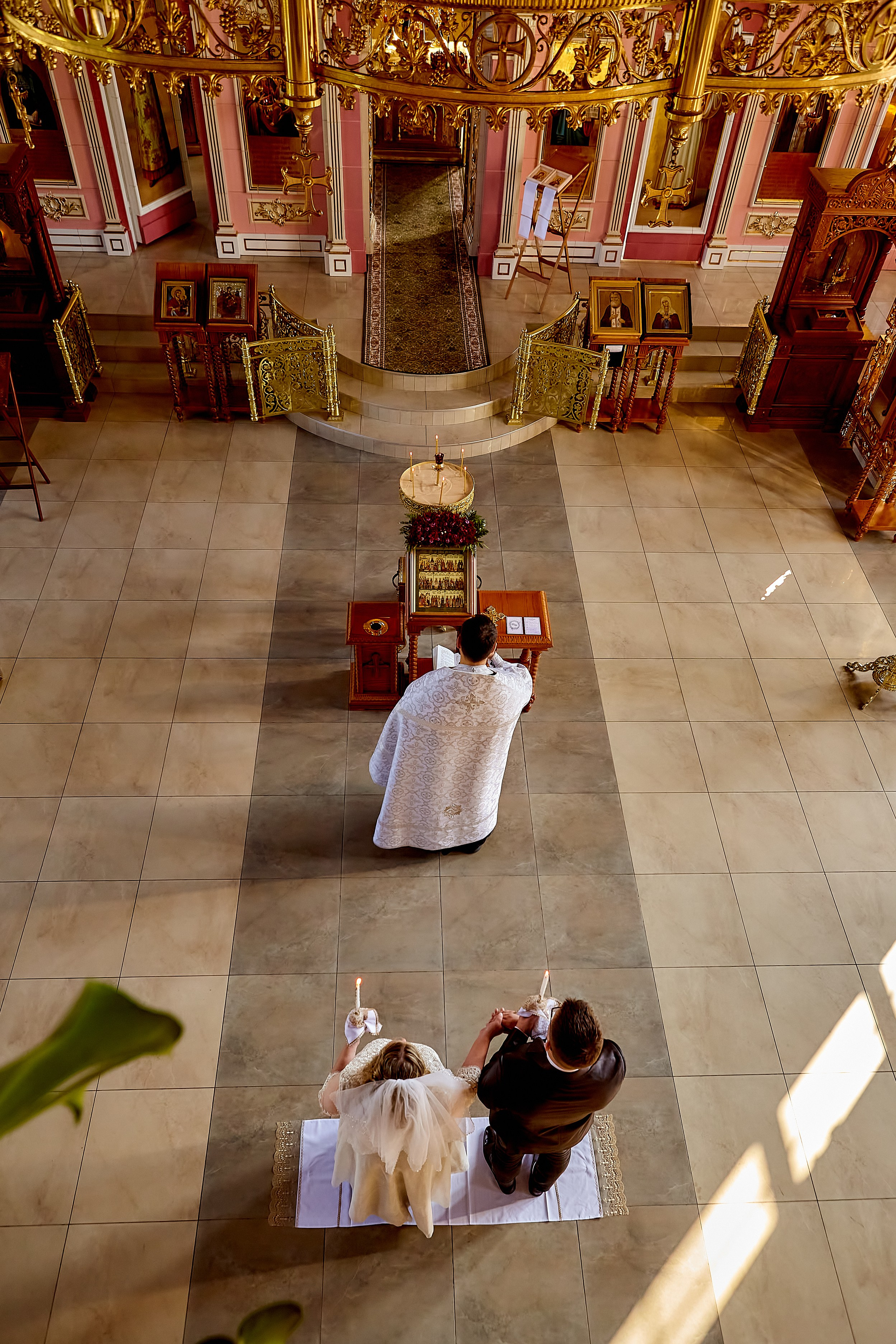 Вид сверху в храме на таинстве венчания жених и невеста со свечами за священником лицом к алтарю.