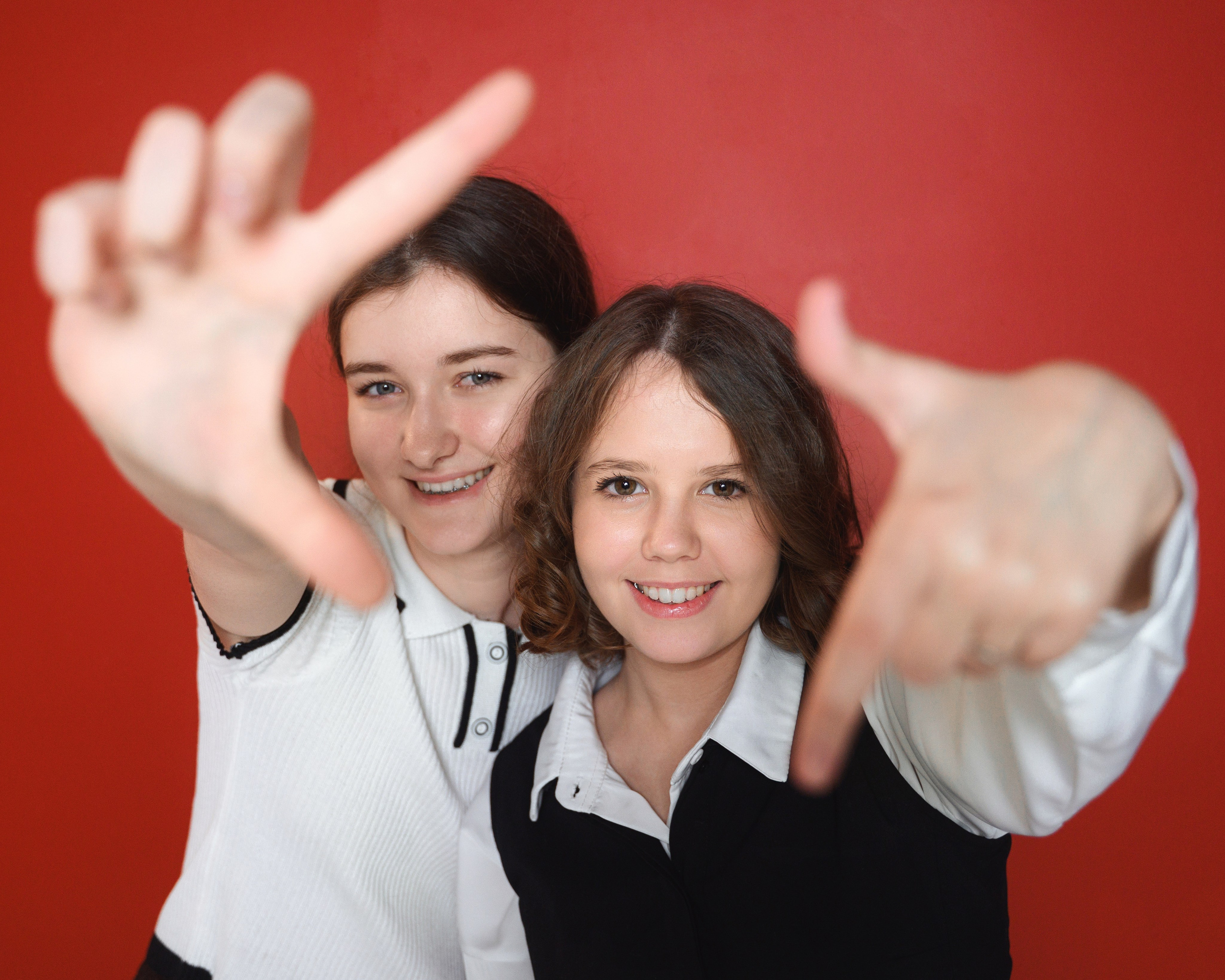 Две ученицы на красном фоне тянут руки к камере в эмоциональном дружеском кадре.