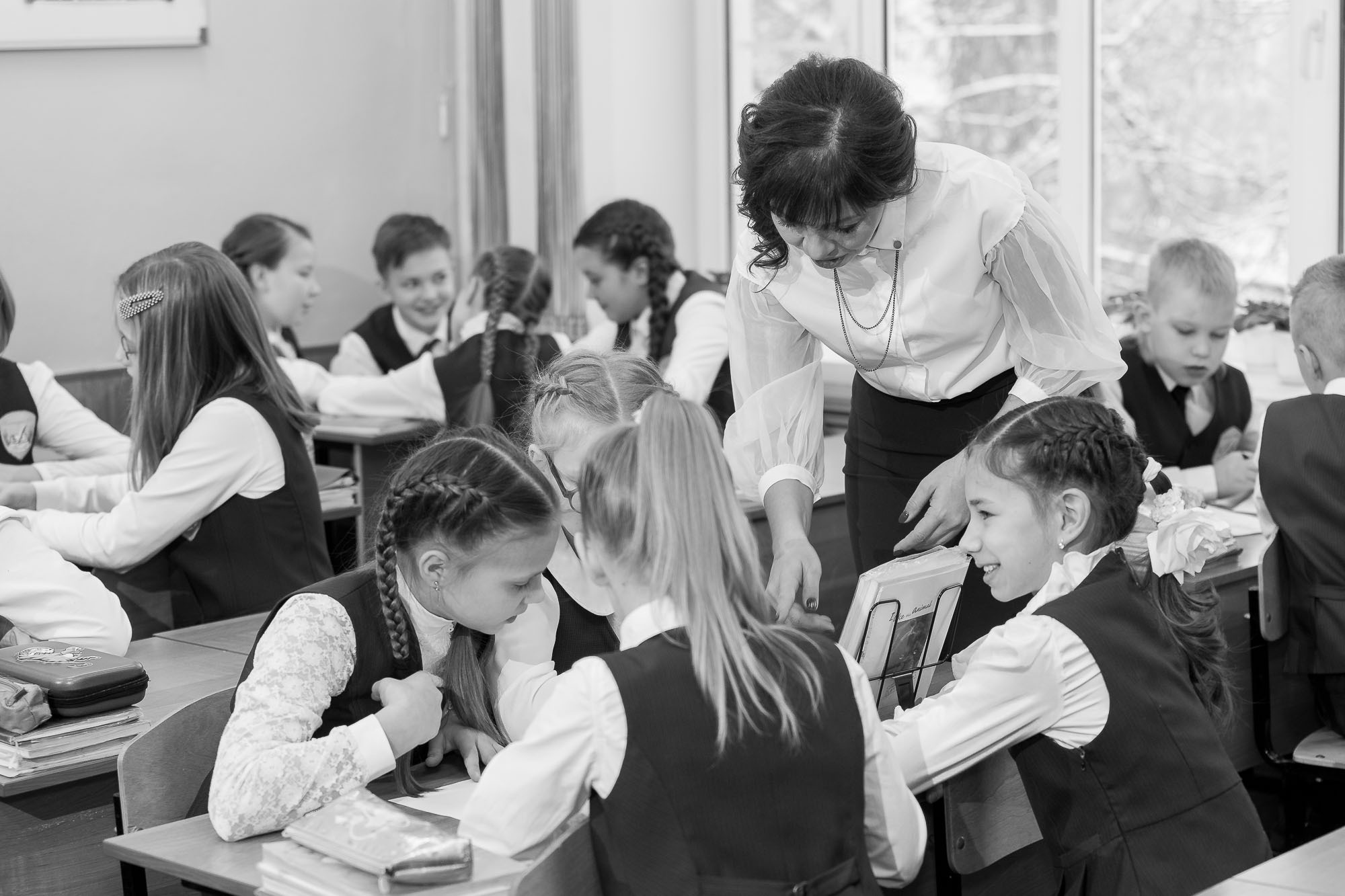 Учитель 4 класса склонился над группой учеников в классе, которые сидят за партой.