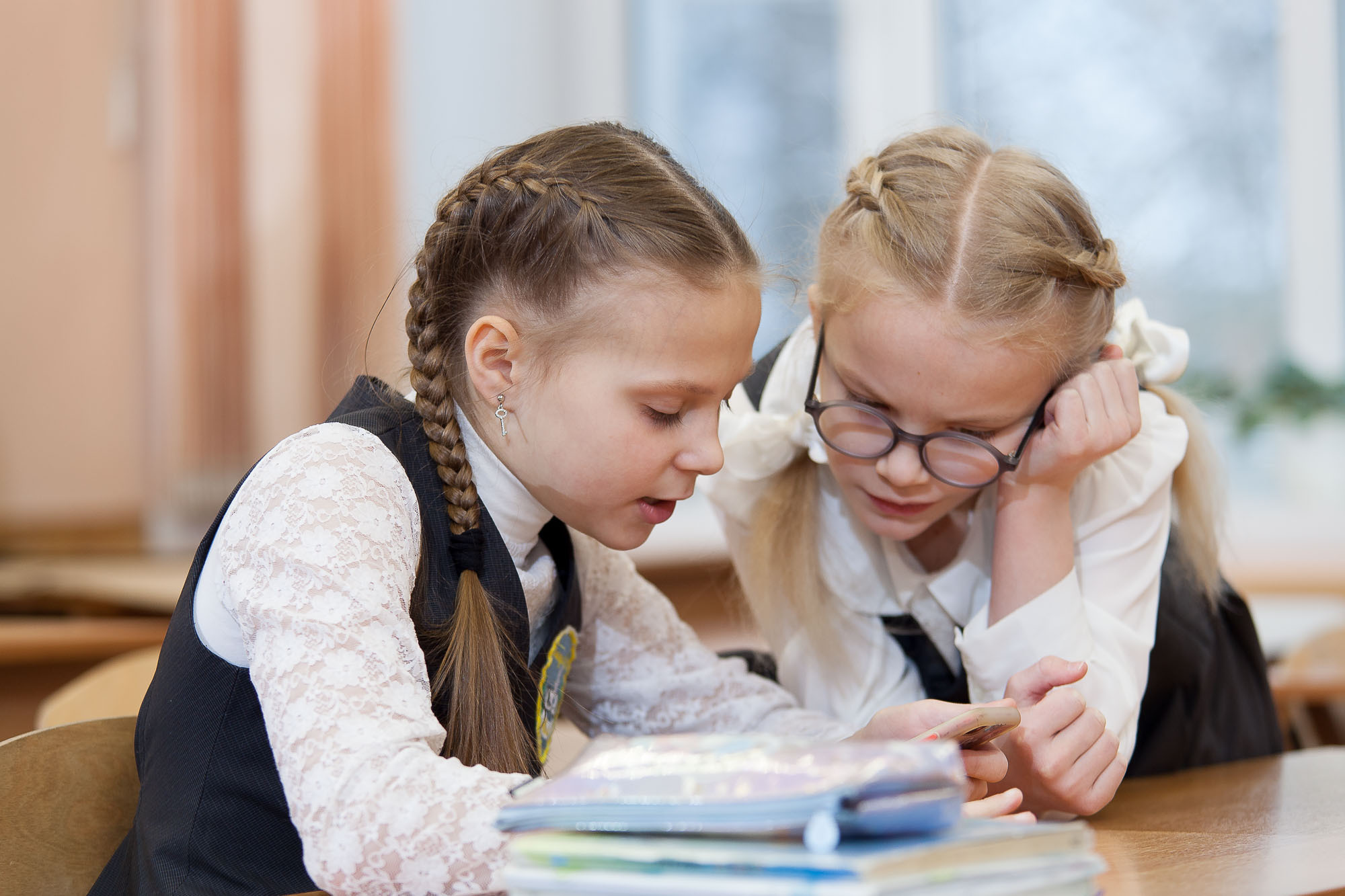Две девочки в черно-белой форме склонились за партой над учебником.