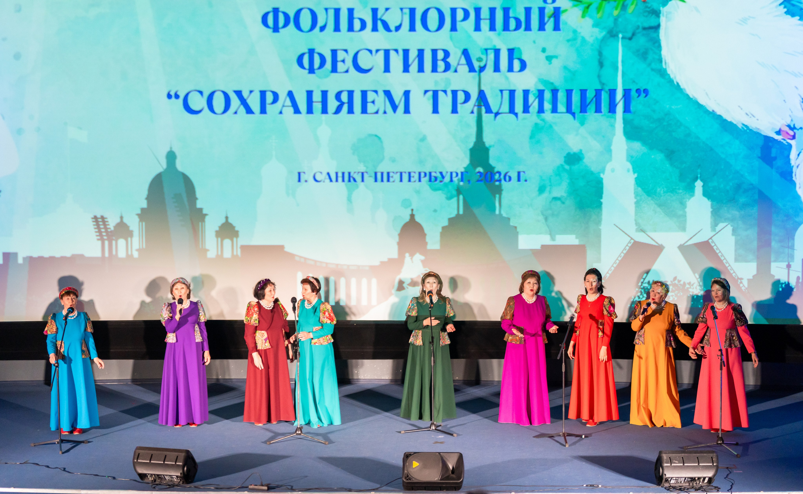 Женский вокальный ансамбль в ярких цветных народных костюмах исполняет песни на сцене XII Международного фольклорного фестиваля «Сохраняем традиции» в Санкт-Петербурге, 2026. Автор фото: Мальцев Евгений.