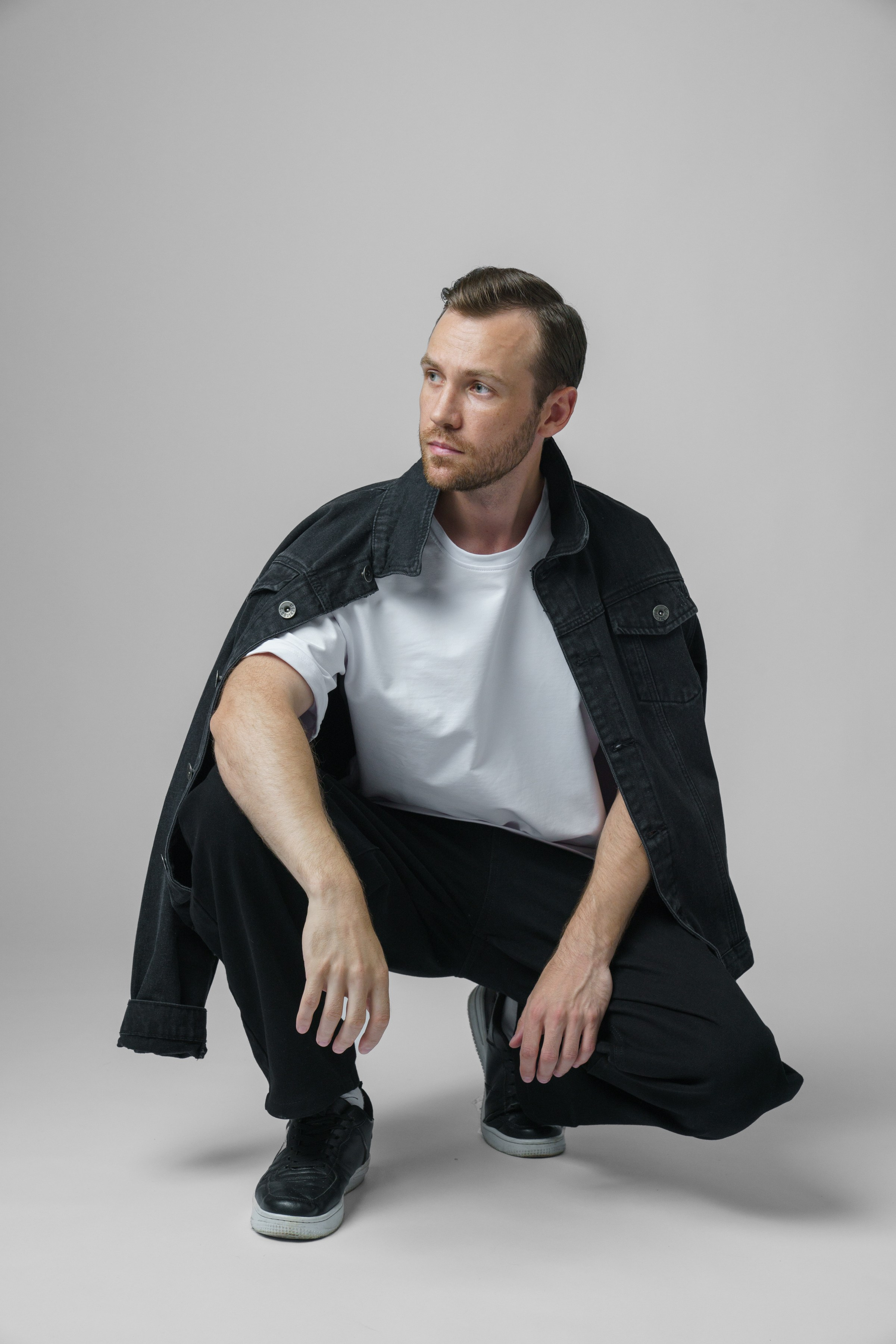 Мужчина с короткой щетиной с накинутой джинсовой курткой на плечи сидит на корточках