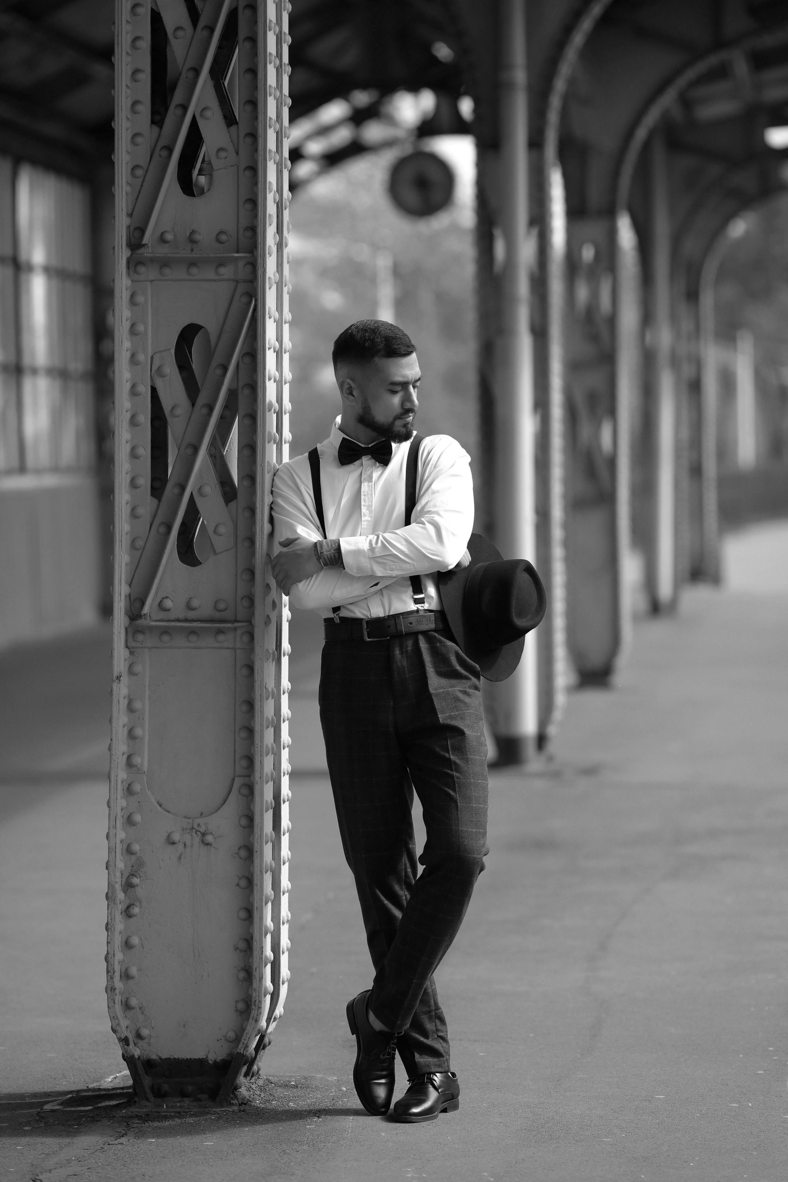 Мужчина с бородой в брюках с подтяжками, белой рубашке с бабочкой стоит со шляпой в руках