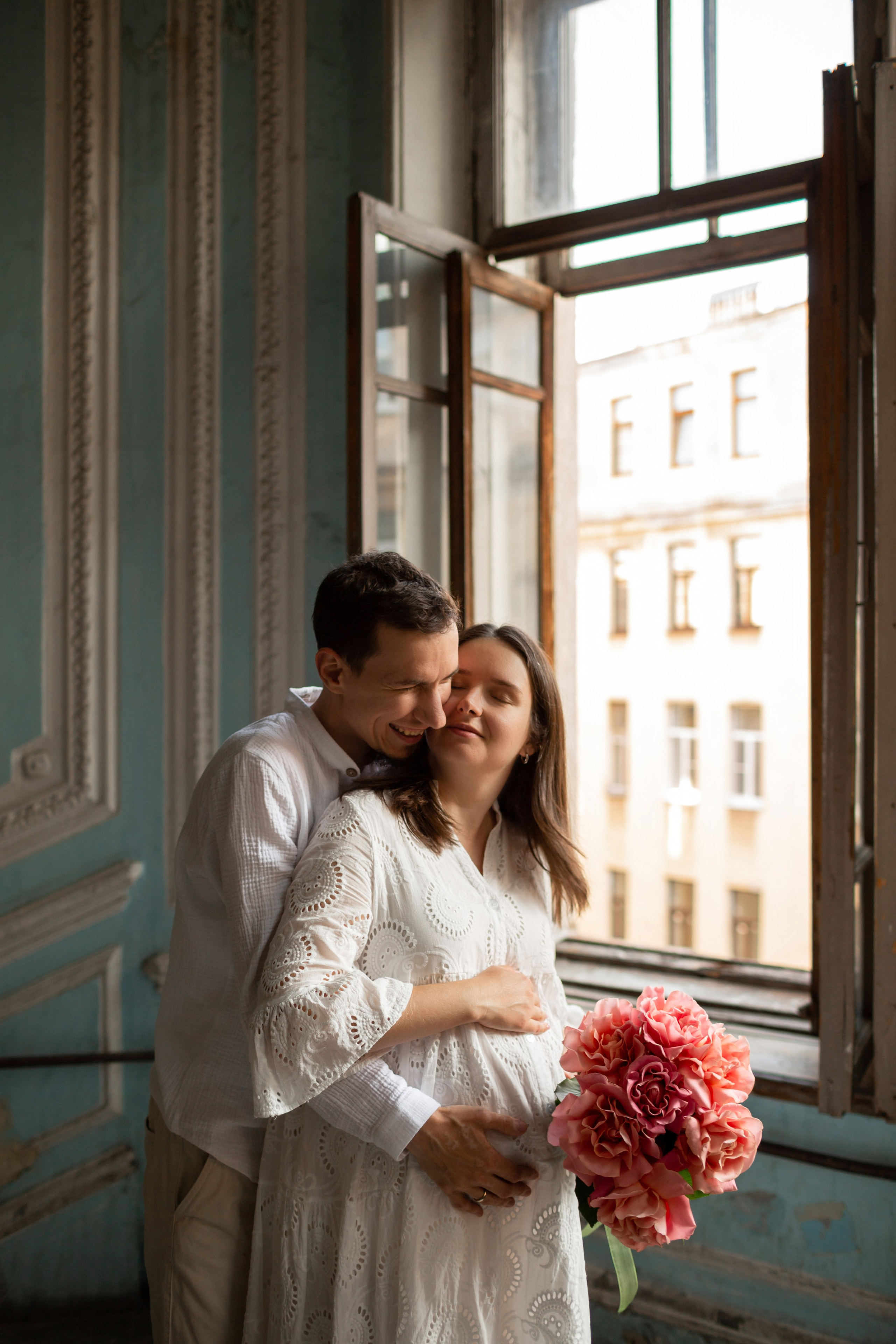 Муж обнимает беременную жену в белом платье с букетом.