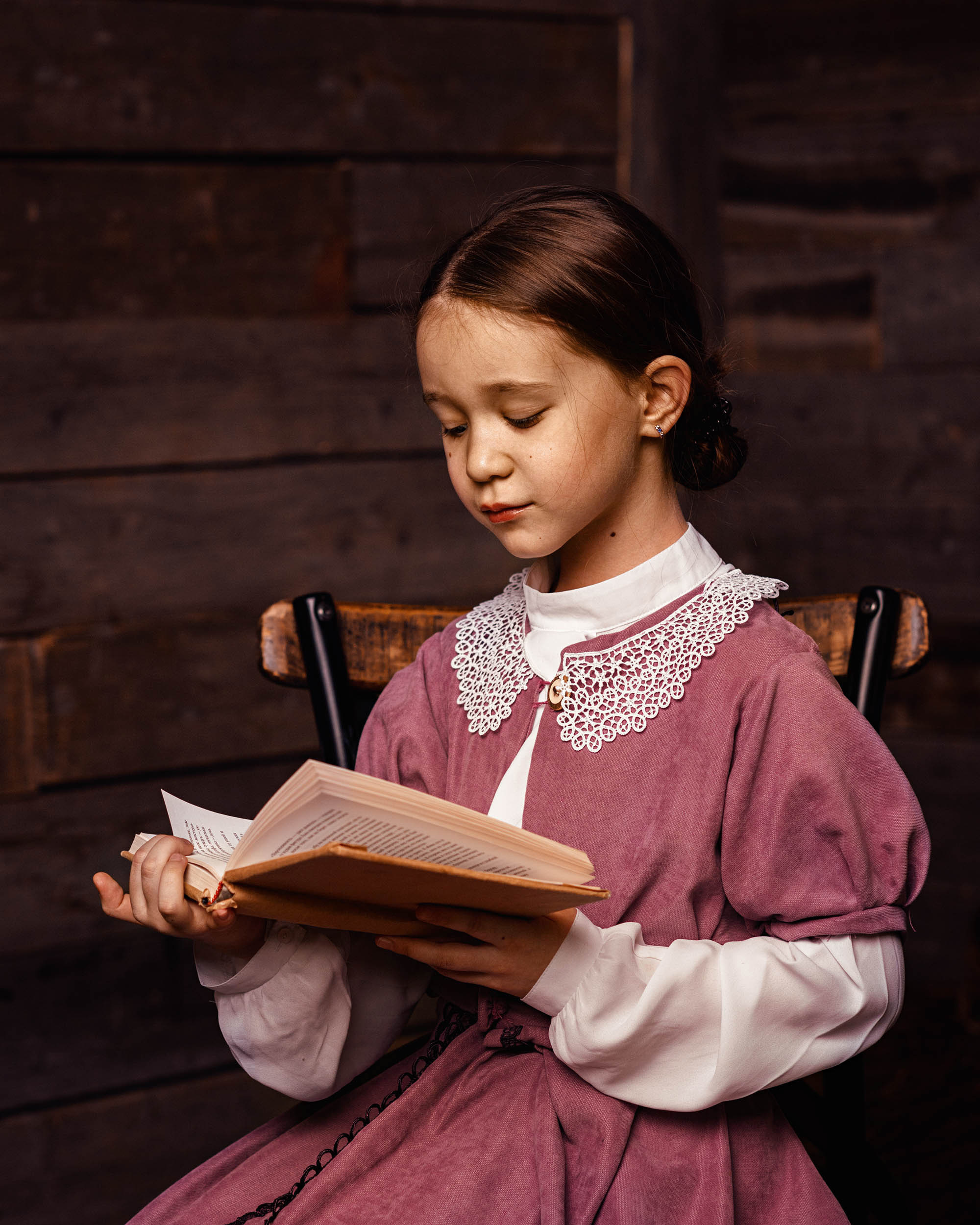 Девочка в красном старинном платье сидя на стуле читает книгу на фоне стены                             