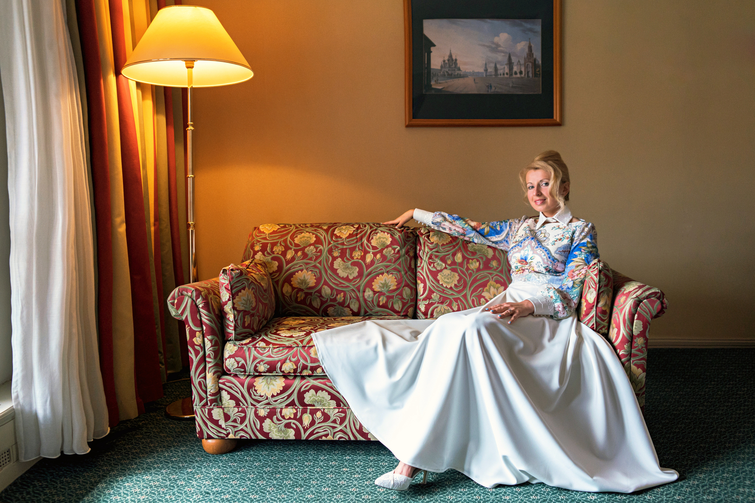 Невеста в необычном платье сидит на диване около окна и высокой лампы