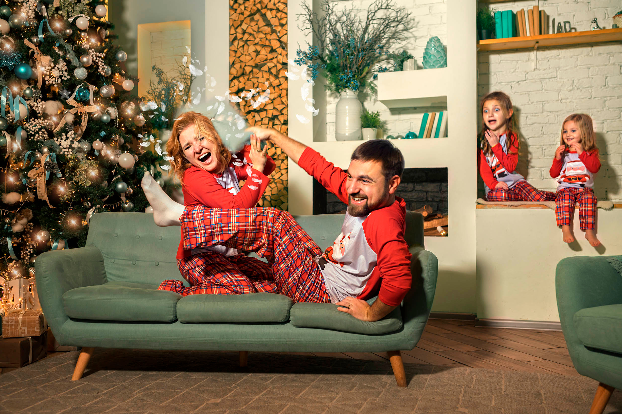Мама в пижаме бьет подушкой папу на диване на фоне новогодней елки и камина, дети за этим наблюдают