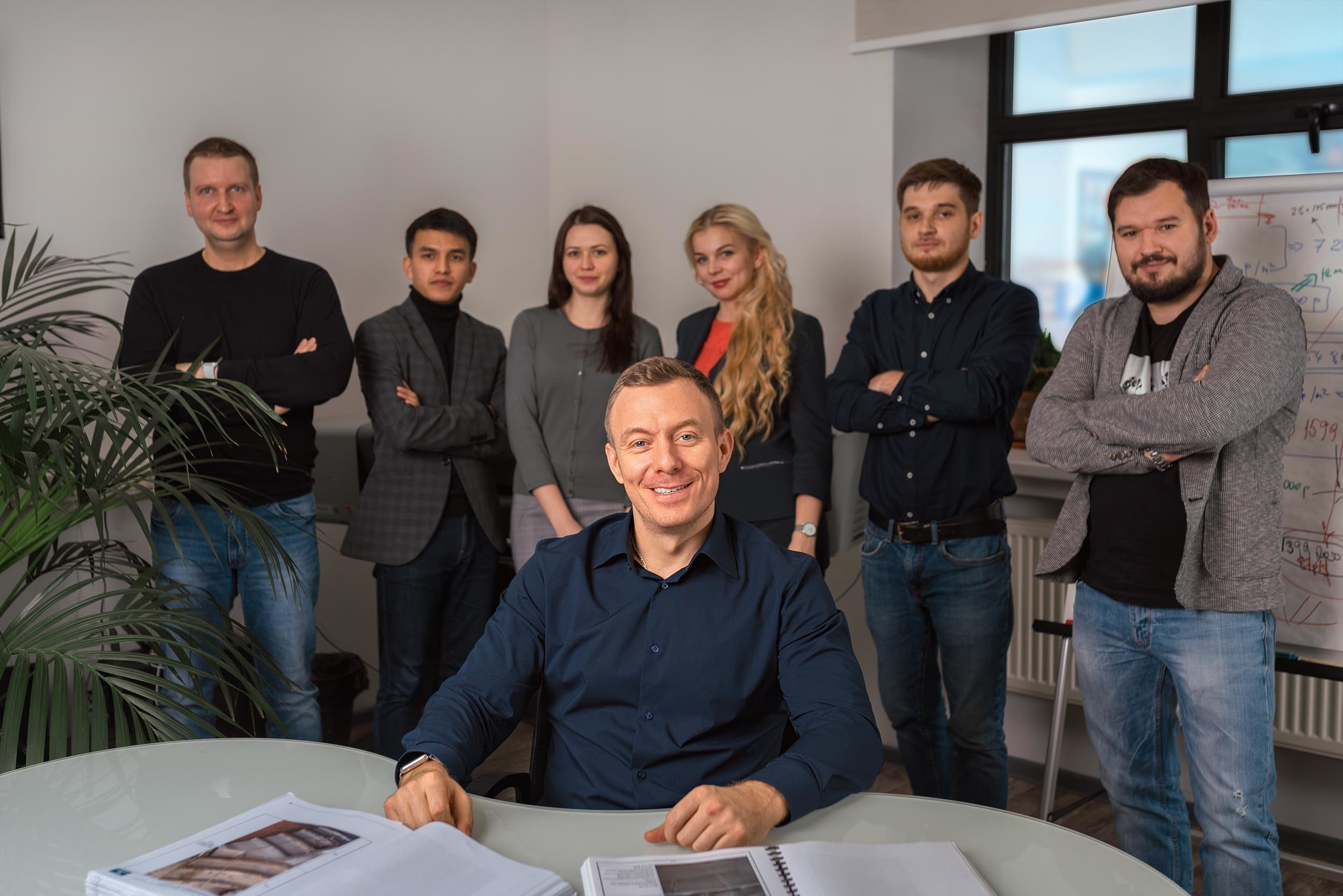 Сотрудники компании смотрят в камеру стоя за столом в офисе, на переднем плане, за столом ситит руководитель