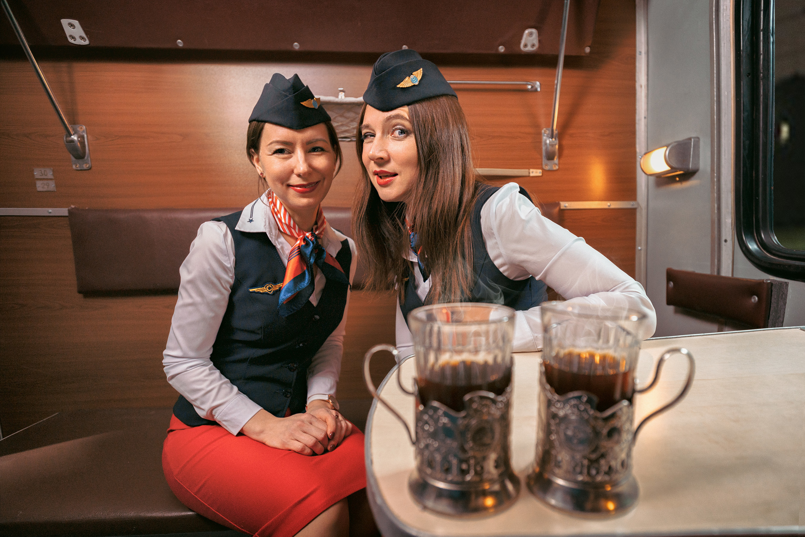 Две проводницы в красных юбках и пилотках в купе поезда назаднем плане, на переднем чашки с чаем