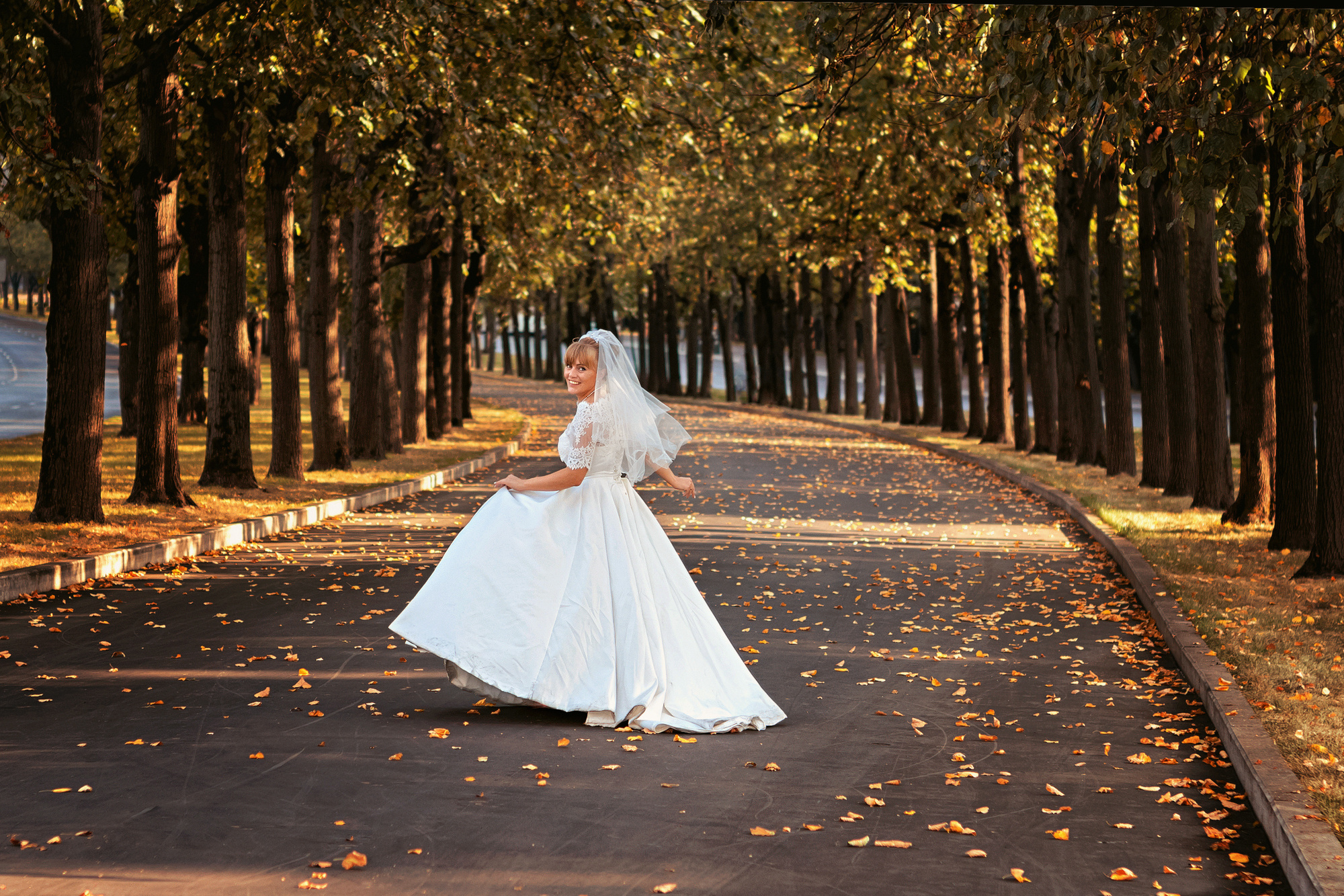Невеста в белом платье и фате бежит по аллее между деревьев