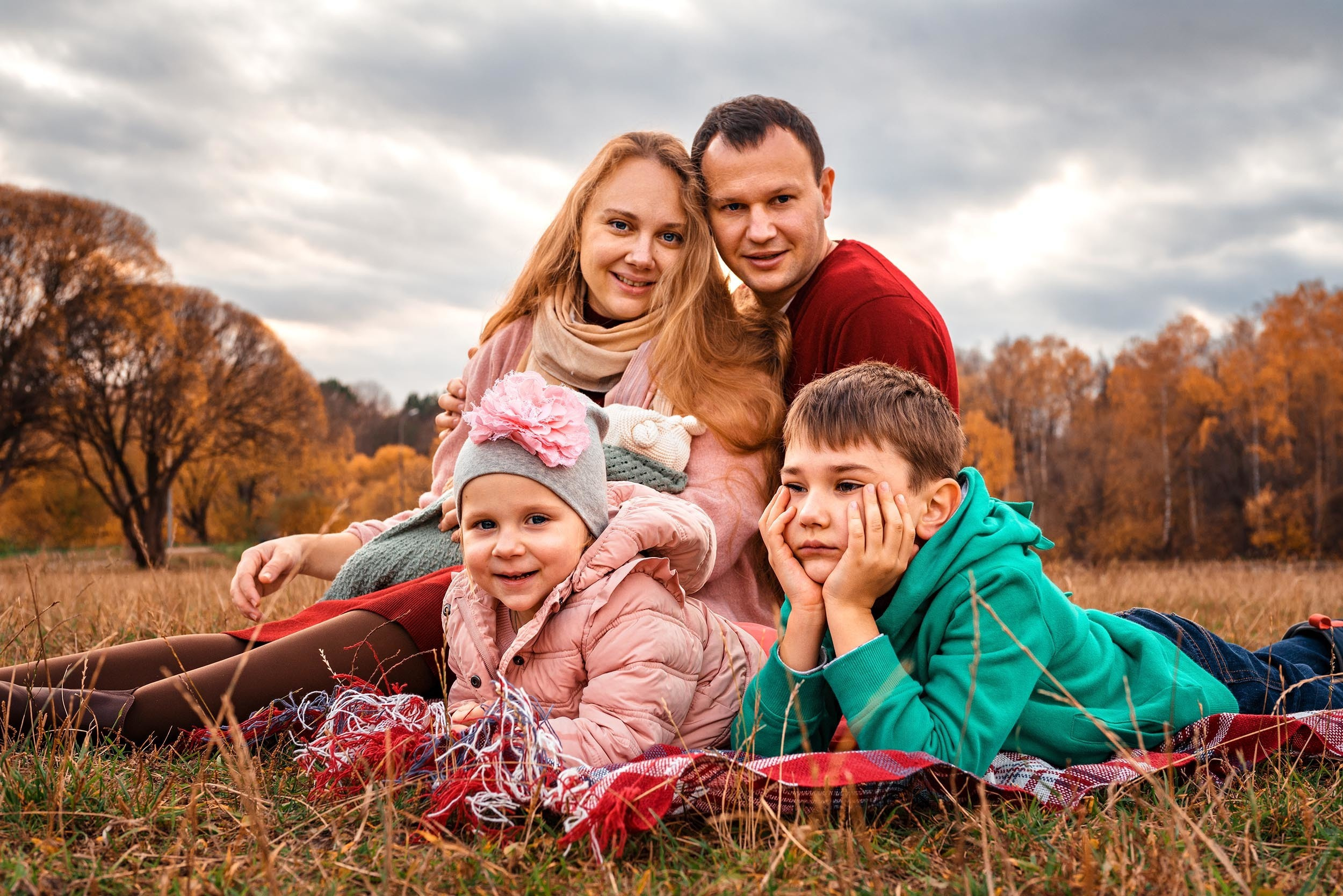 Семья смотрят в камеру на пледе посреди поляны на фоне осеннего леса и облаков