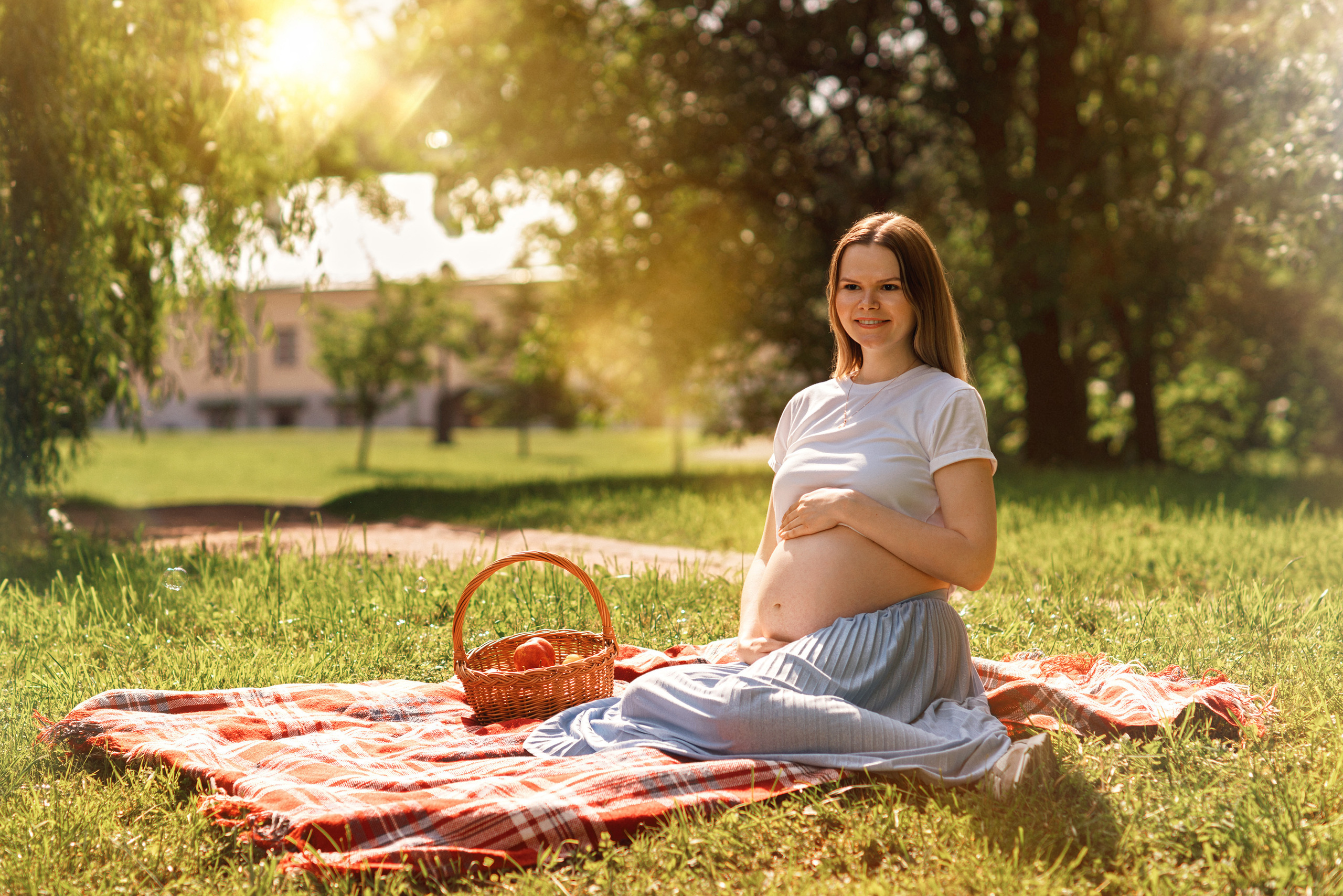 Беременная девушка сидит с корзинкой на пледе в парке