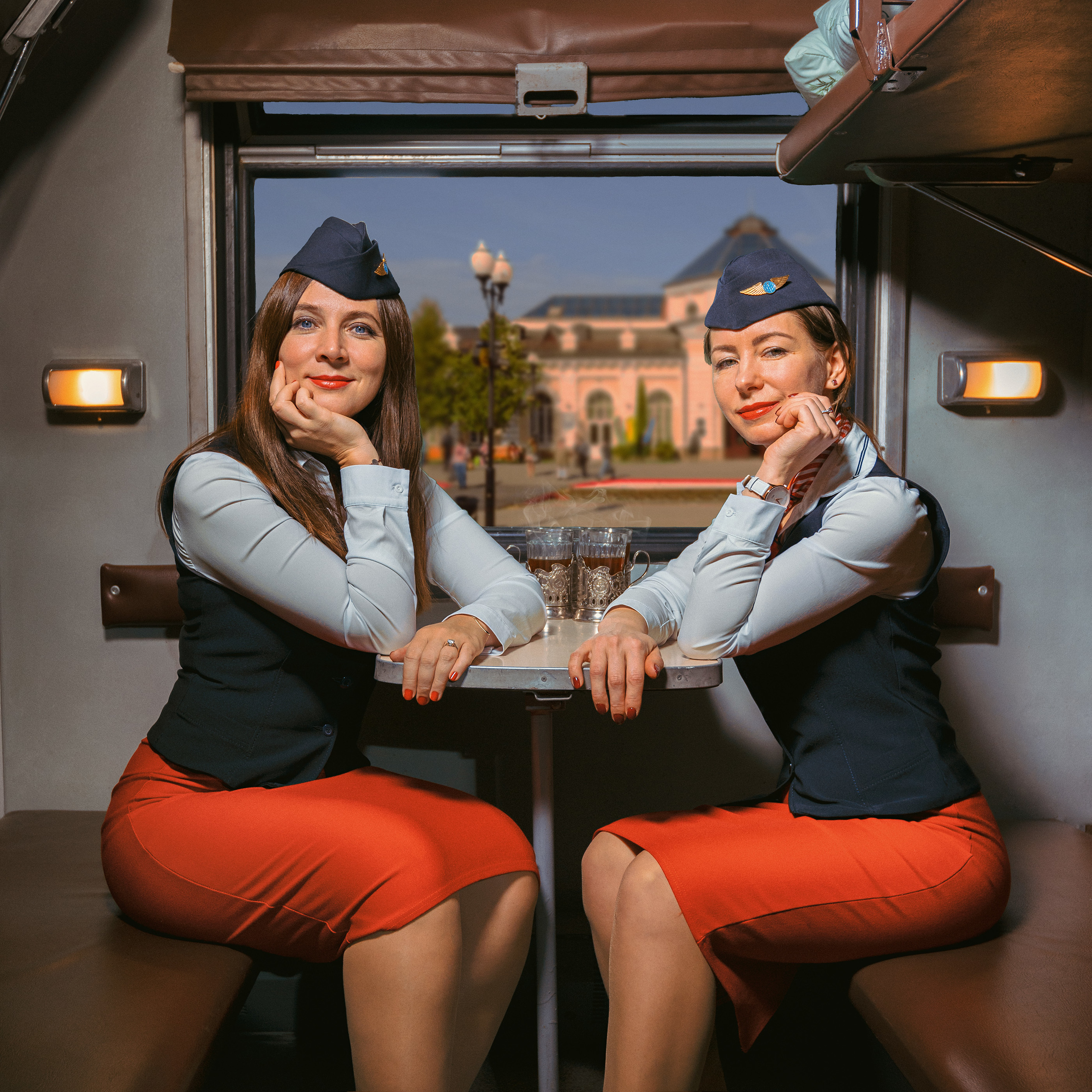 Две проводницы в красных юбках и пилотках сидят в купе поезда на фоне окна