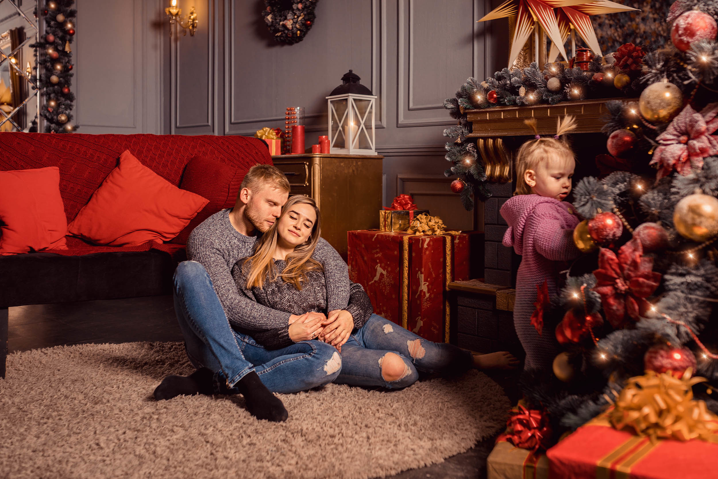 Родители обнимаются сидя на полу, дочь стоит у камина и наряженой елки