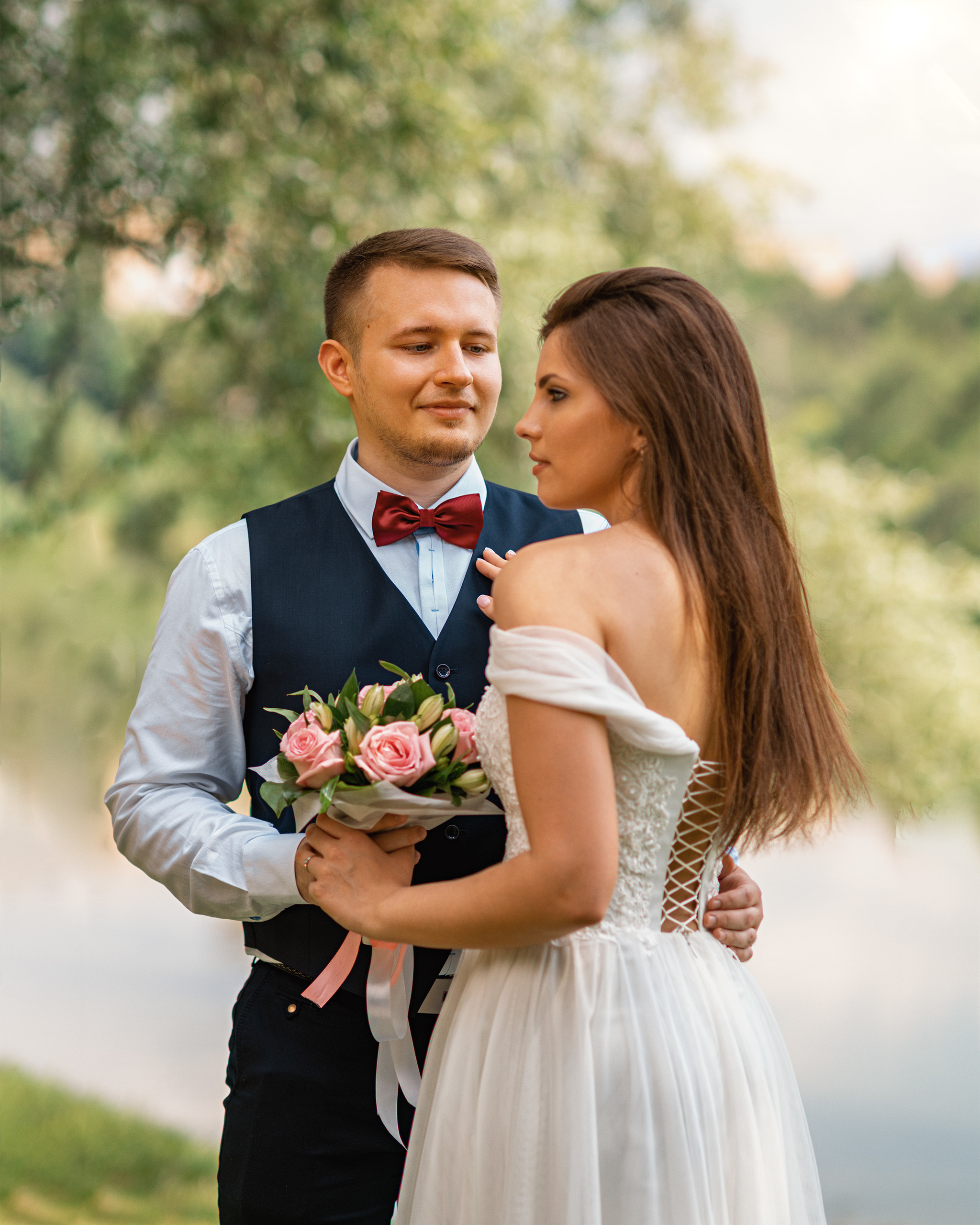 Жених смотрит на невесту, оба держастя за букет, у невесты красивая шнуровка платья на спине
