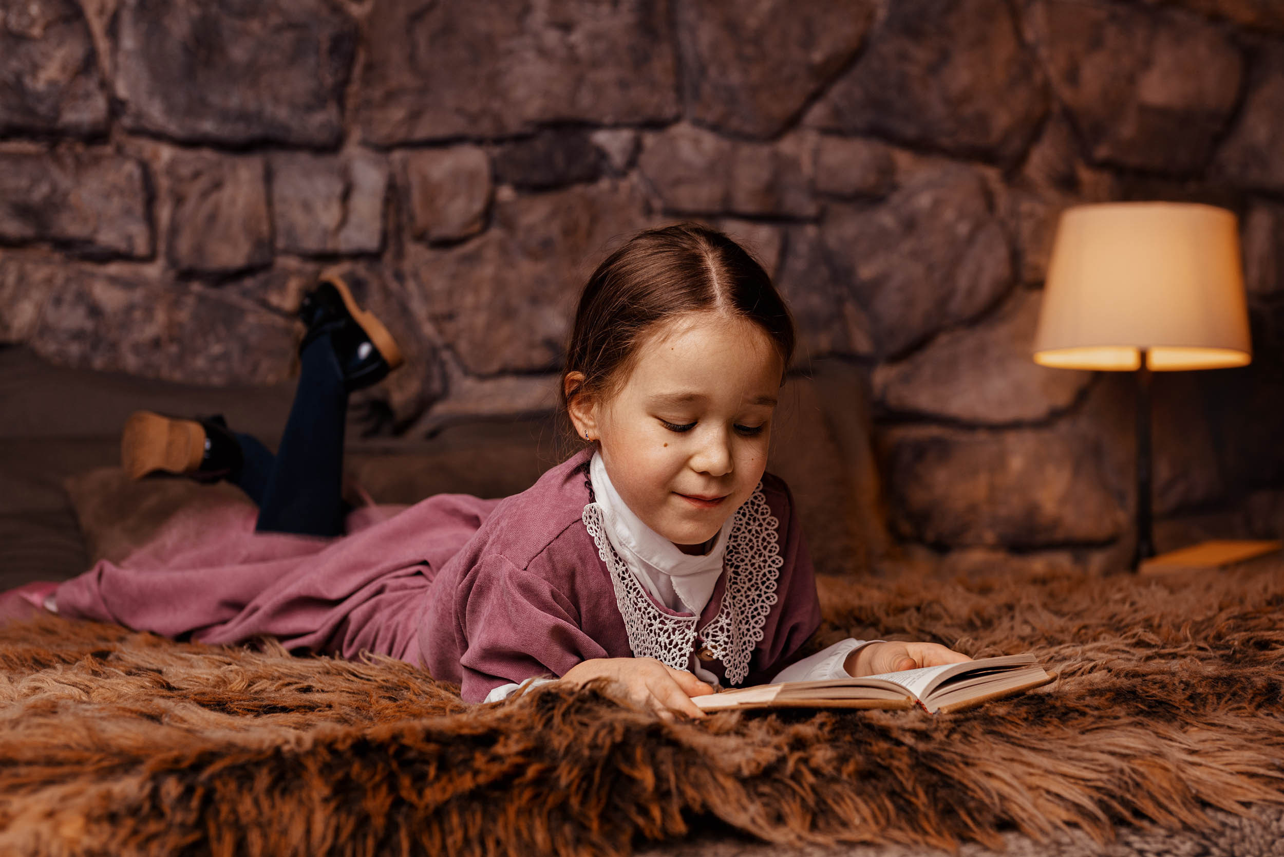 Девочка в красном старинном платье читает книгу лежа на шерстяном пледе на фоне каменной стены и лампы