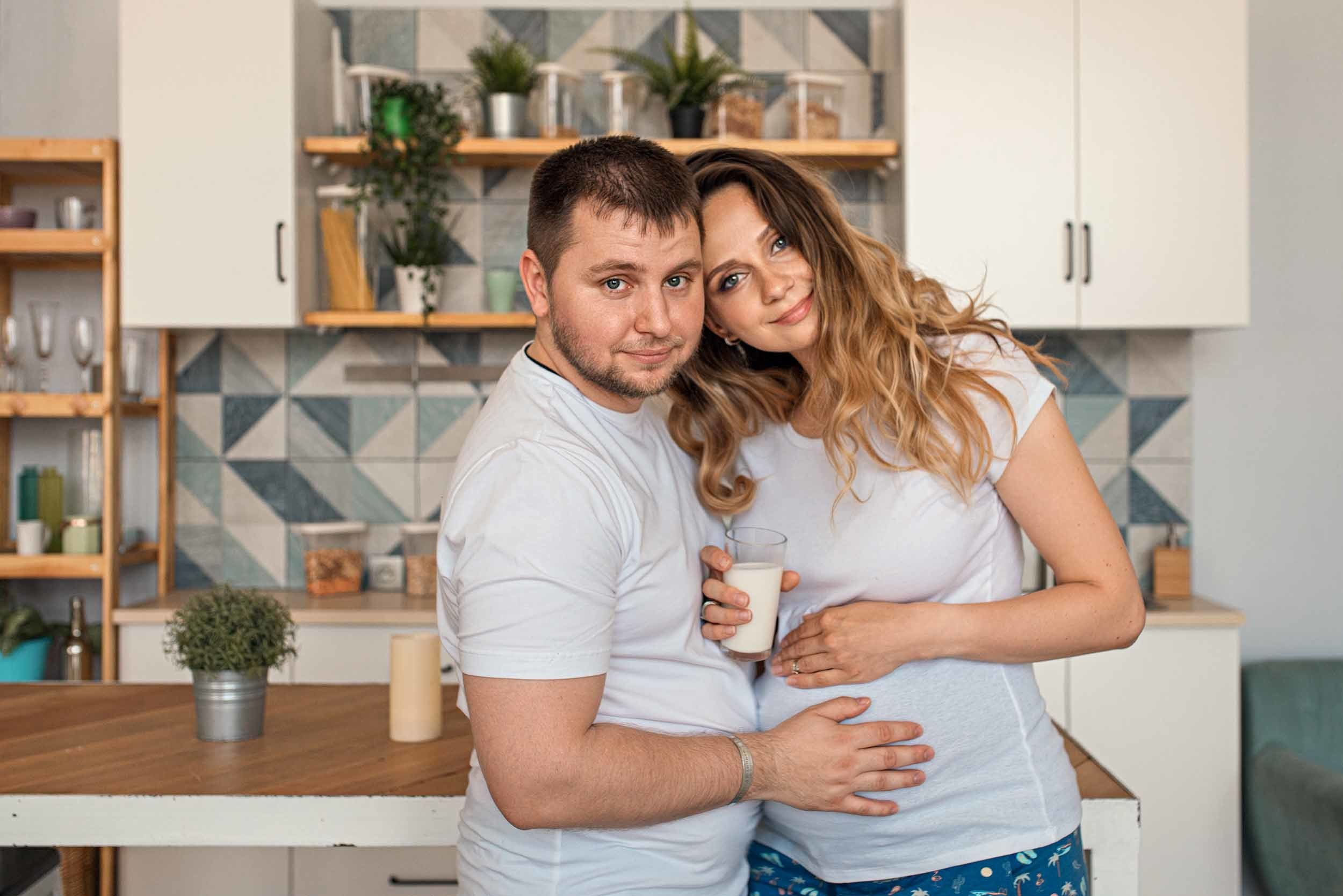 Беременная девушка и мужчина обнимаясь смотрят в камеру стоя на кухне