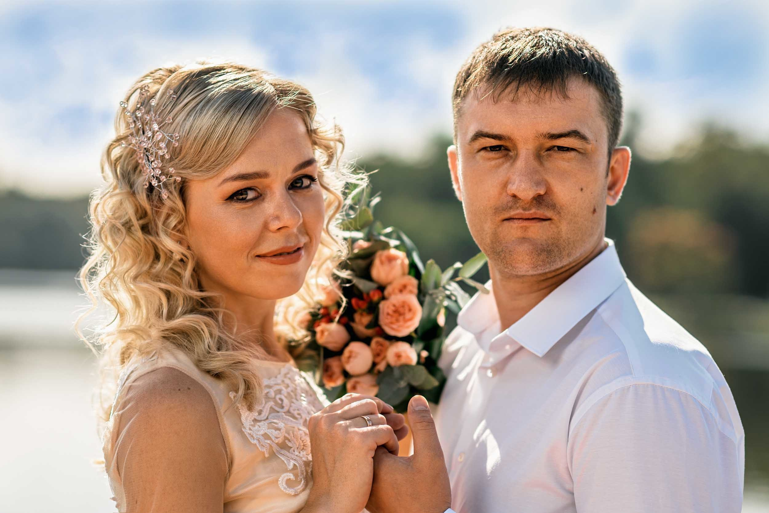 Пара молодоженов держатся за руки и букет, смотрят в камеру на берегу