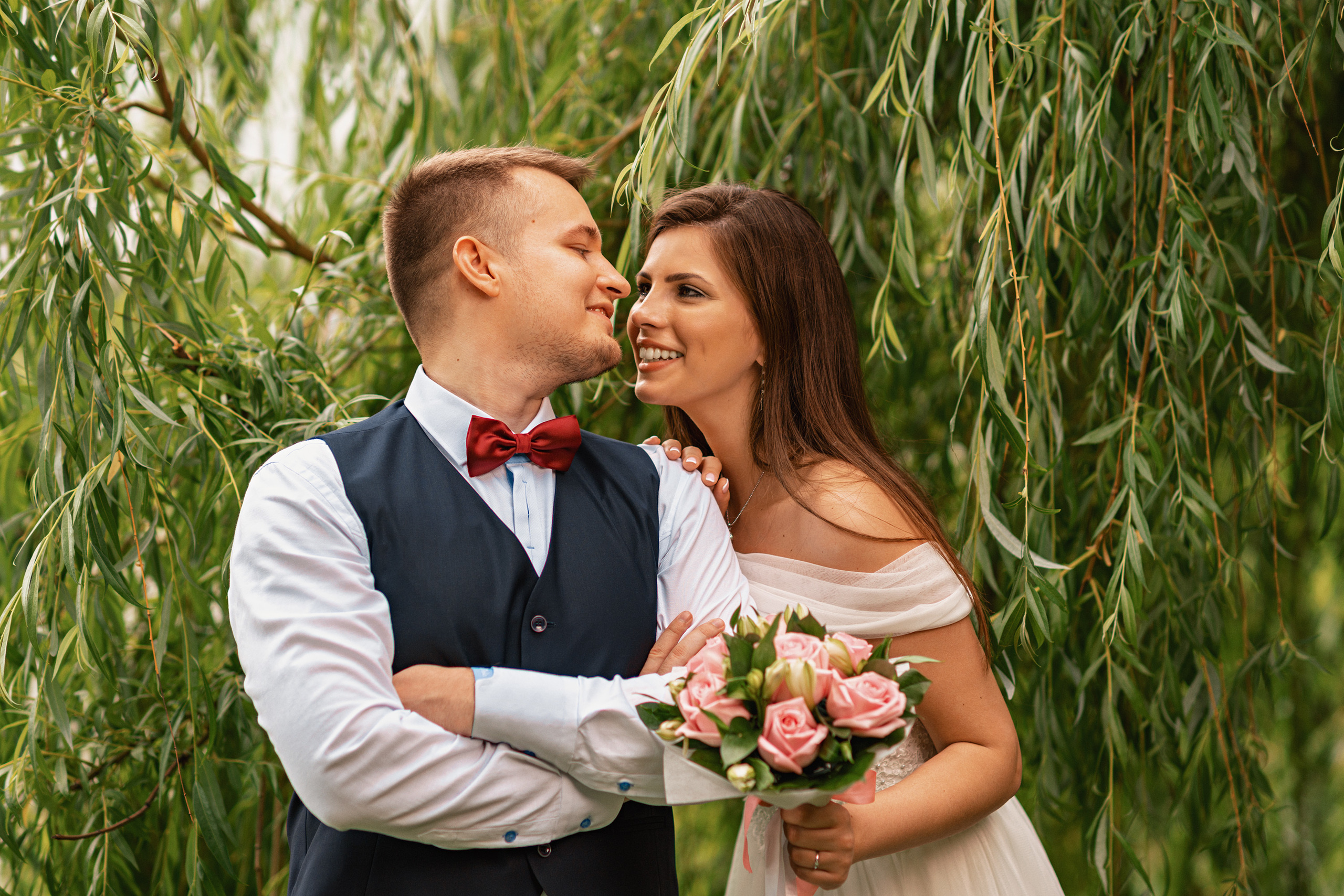 Жених и невеста смотрят друг на друга, у невесты в руке букет, сзади листья