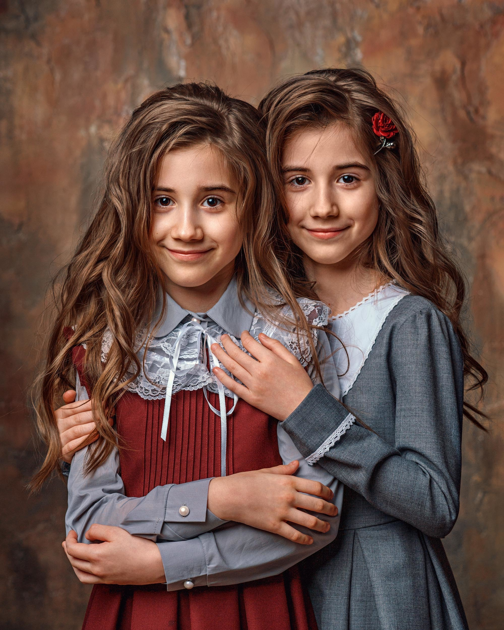 Две сестры, двойняшки с длинными волосами в красивых платьях обнимаясь смотрят на зрителя