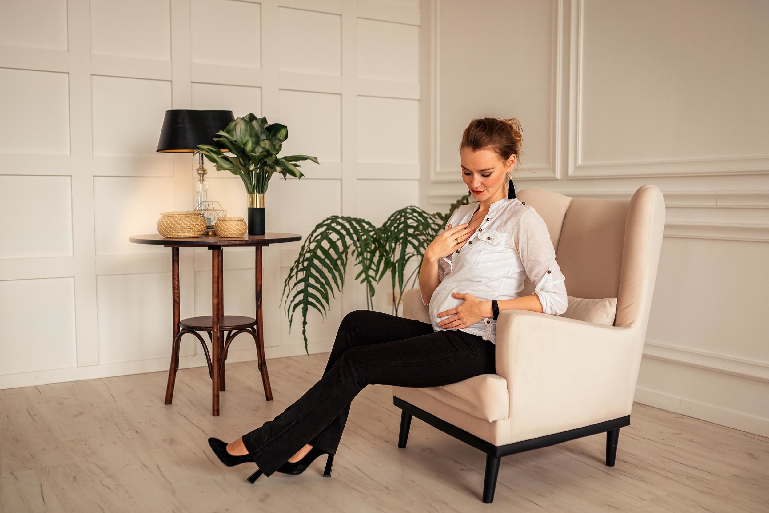 Беременная девушка смотрит на живот сидя в кресле посреди белой комнаты, рядом стол с лампой и цветами