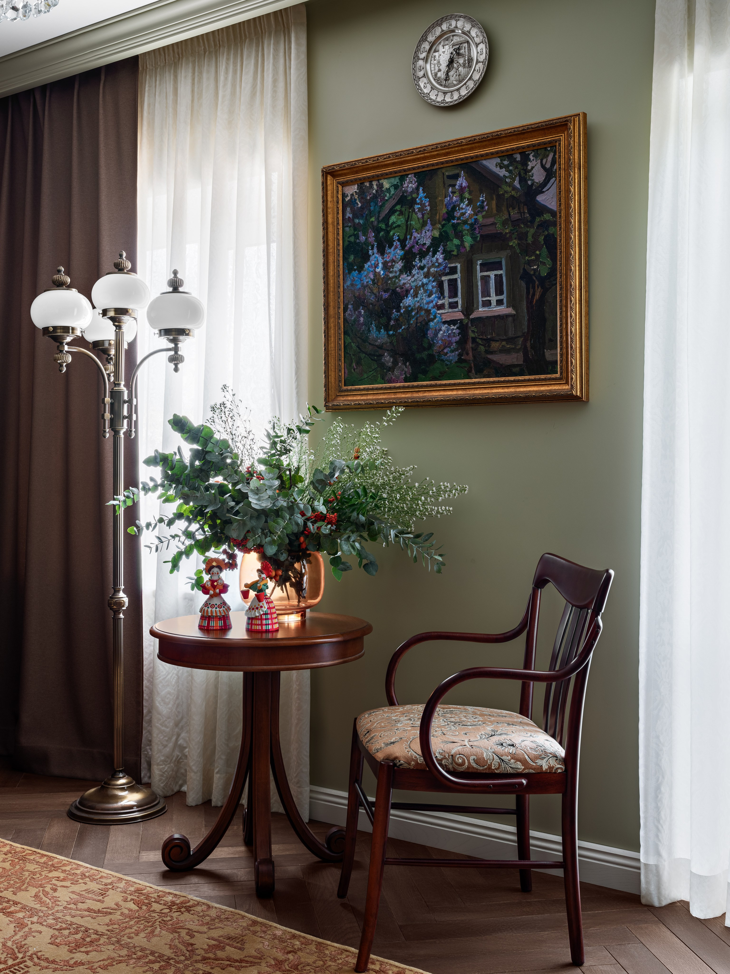 Уютный уголок гостиной с торшером в классическом стиле, круглым деревянным столиком, букетом зелени и картиной в золотой раме на фоне мягких штор; интерьер гармонирует теплом и атмосферой. Дизайнеры Юлия Капункина и Альбина Терещенко, стилист Алина Нелень, фотограф Сергей Денисенко.