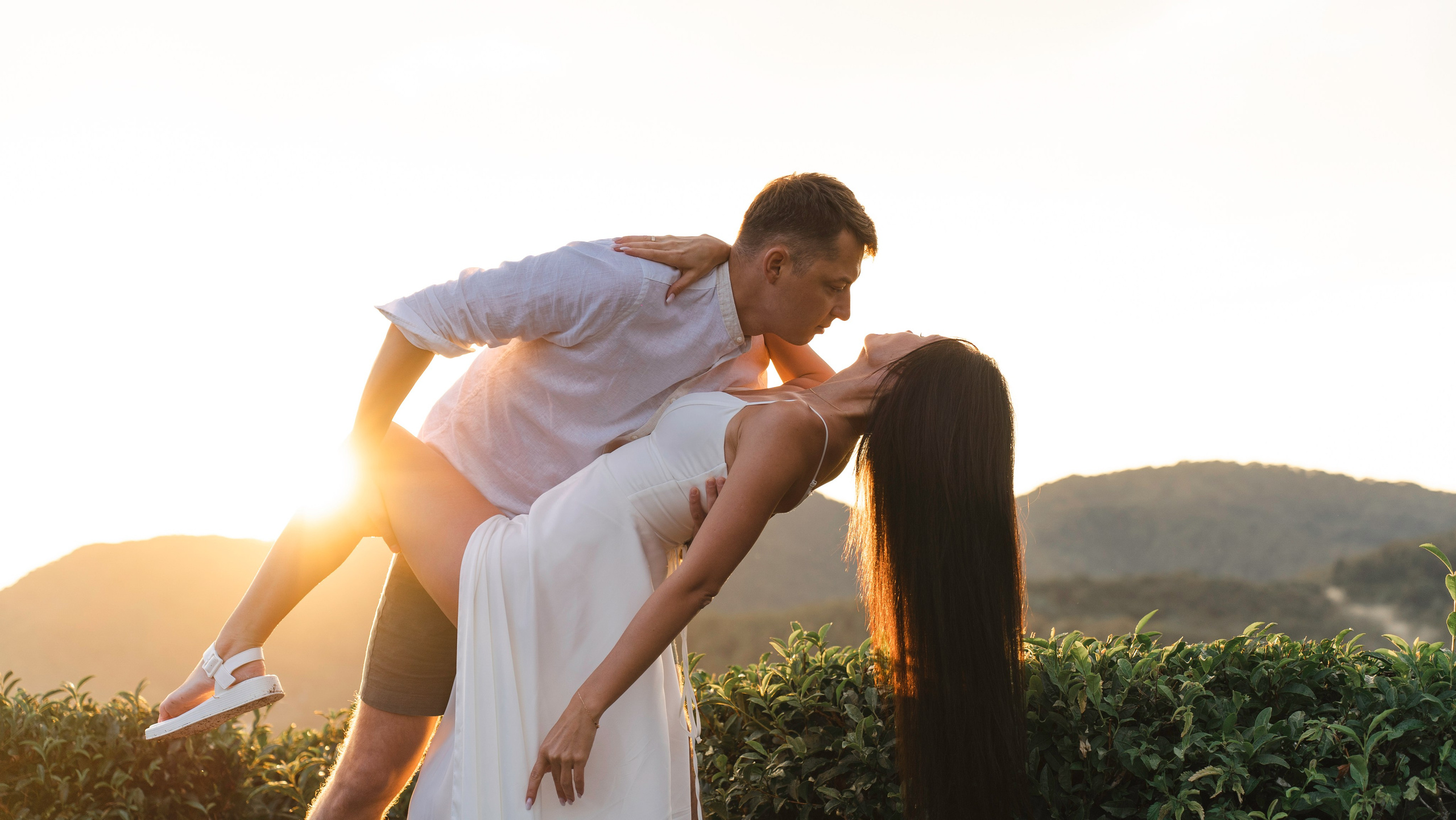 На этой фотографии изображена пара, занимающаяся игривыми и нежными моментами на улице во время заката. Фон включает в себя пышную зелень и холмы. Женщина в белом платье стоит, в то время как мужчина, одетый в светлую рубашку и шорты, наклоняется к ней. Теплый, золотистый свет заката добавляет романтическую атмосферу этой сцене.