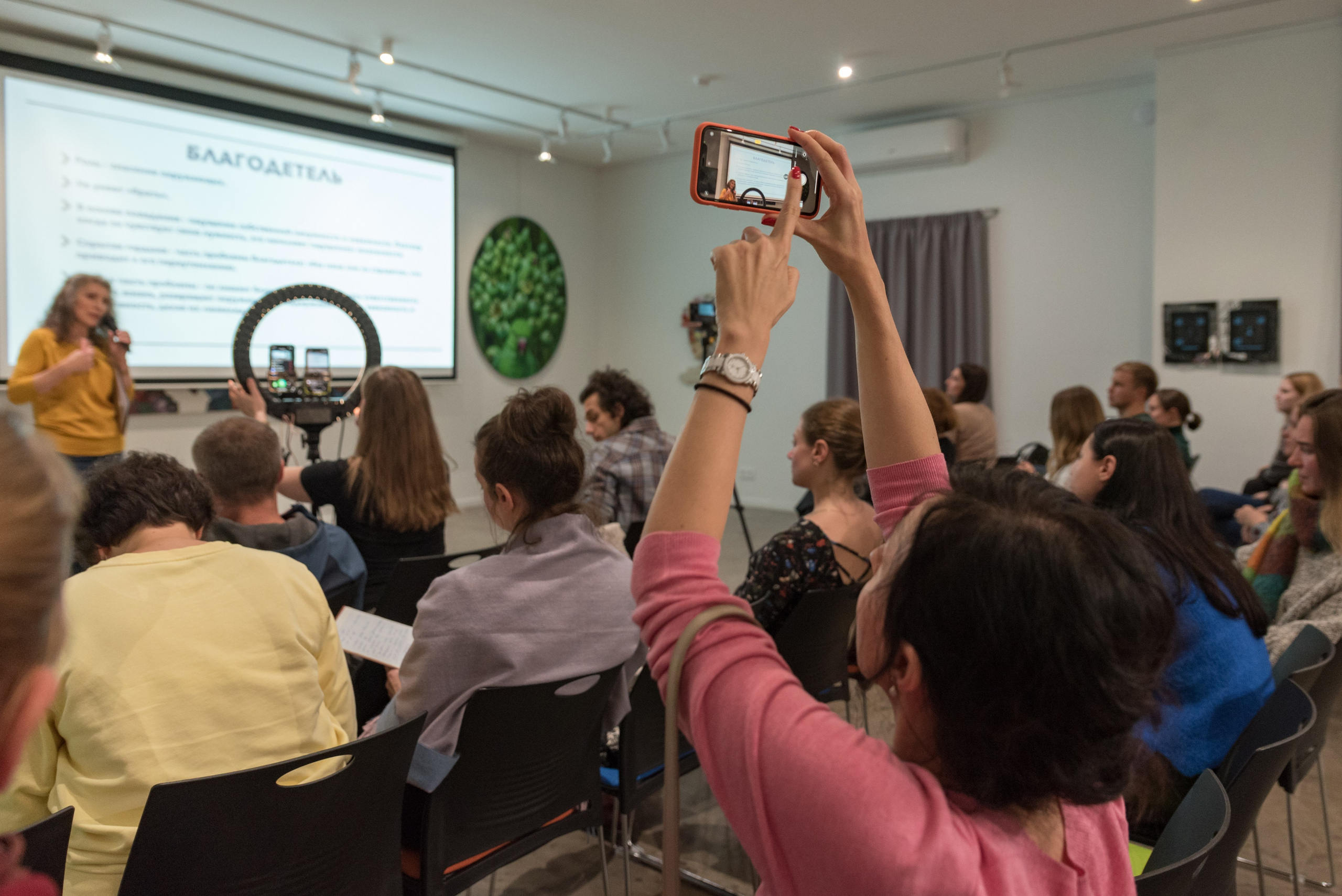 Зрительный зал, женщина снимает лекцию на мобильный телефон поднятый высоко над головой.