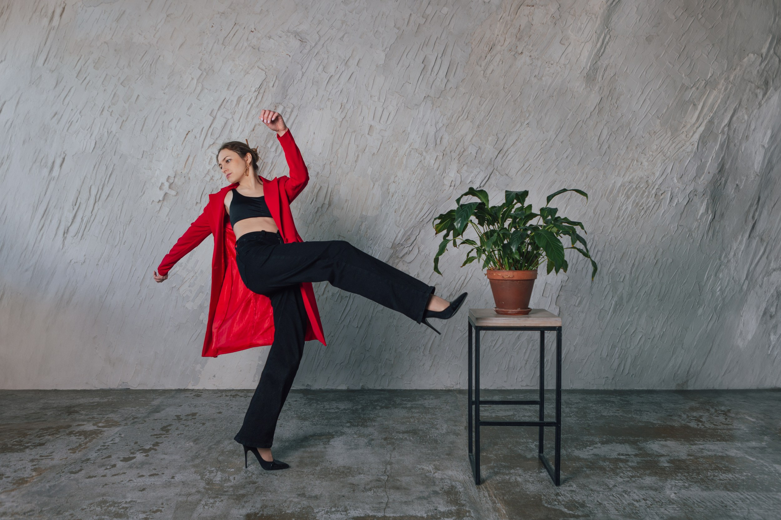 Женщина в красном теряет равновесие на сером бетонном фоне. Зеленое растение с большими листьями в горшке стоит на высокой табуретке.