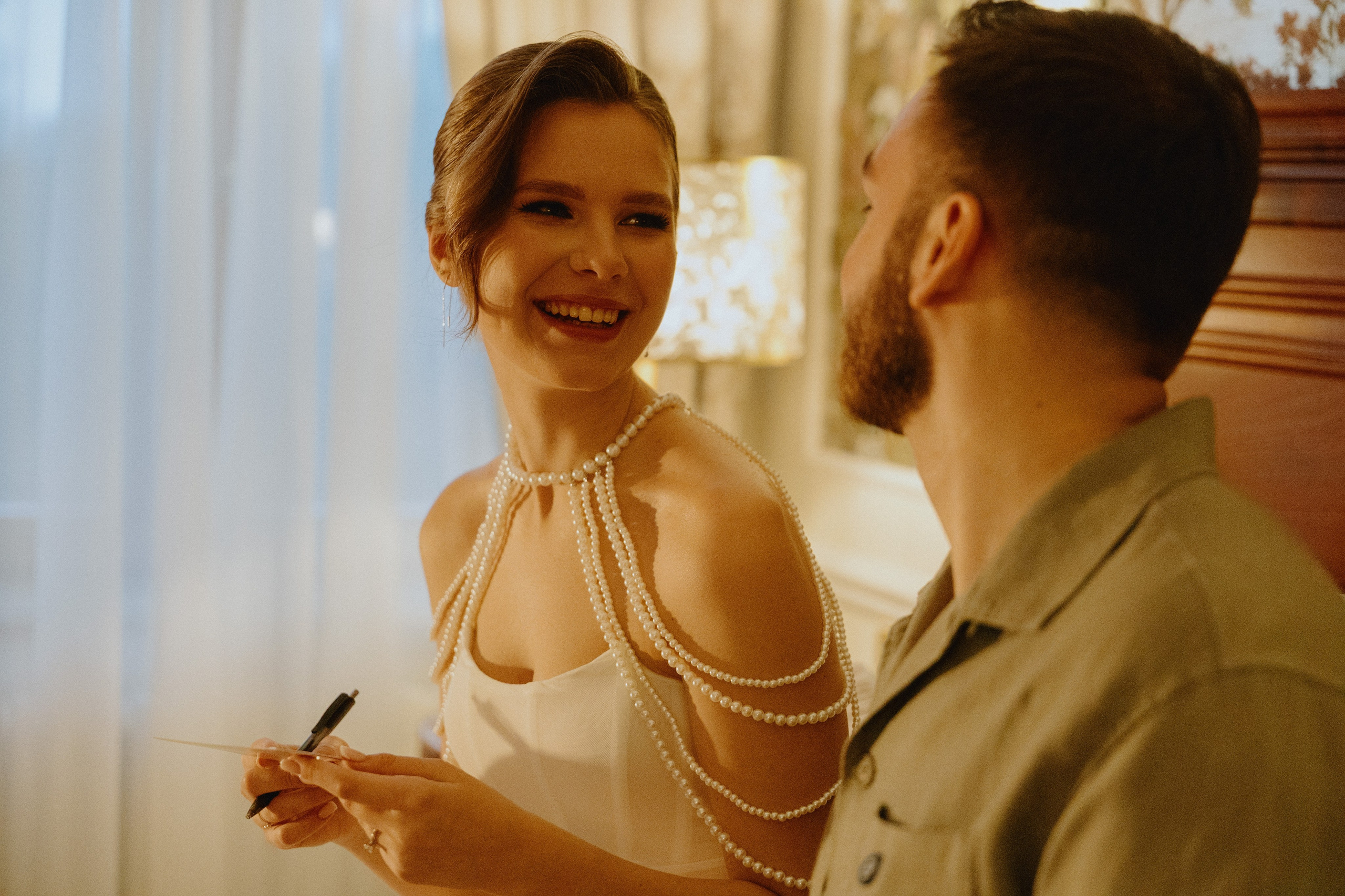 Невеста в свадебном платье с жемчужным ожерельем смотрит на жениха.