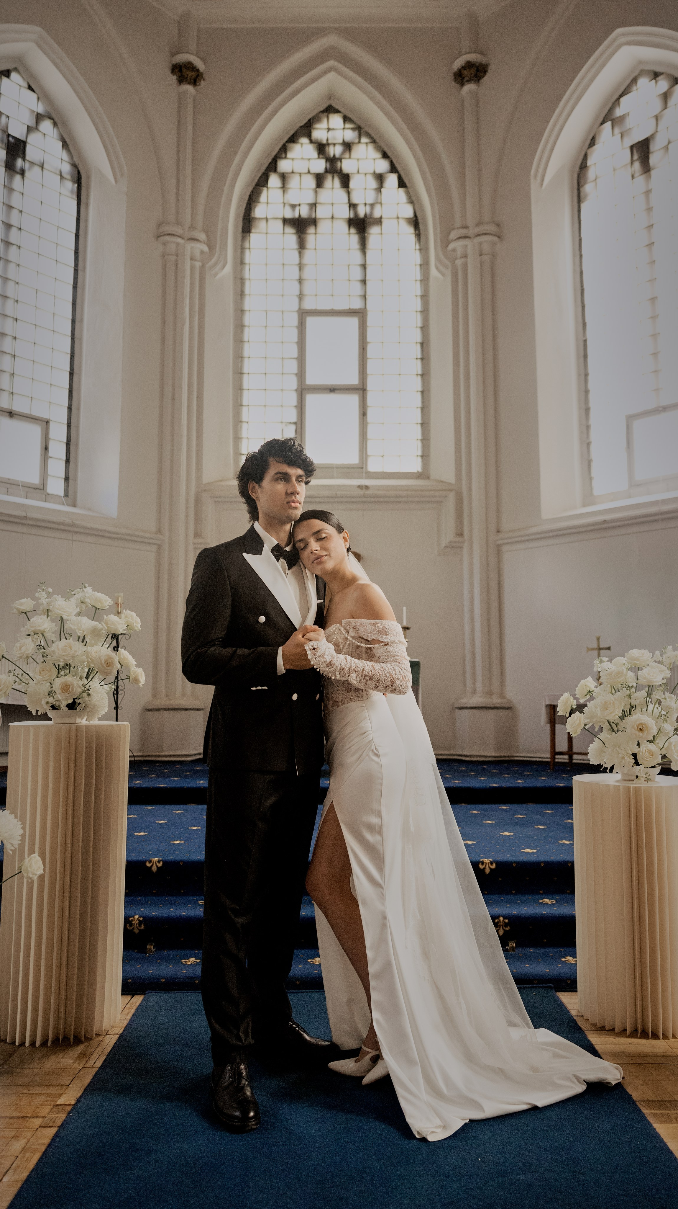 «Свадебный портрет в полный рост в церкви. Жених в чёрном смокинге стоит рядом с невестой в платье с открытыми плечами, длинными кружевными рукавами и высоким разрезом, опираясь на его плечо. Они стоят на синей дорожке, уложенной вдоль прохода, в окружении белых цветочных композиций».