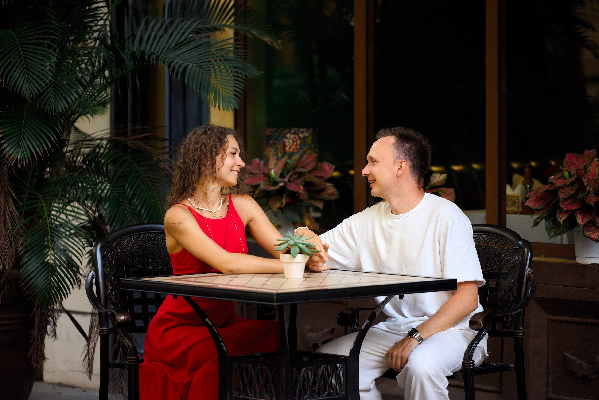A smiling couple in red and white clothes sitting at a black iron outdoor cafe table with a small plant pot between them, surrounded by tropical plants in Sunset Town, Phu Quoc, Vietnam.