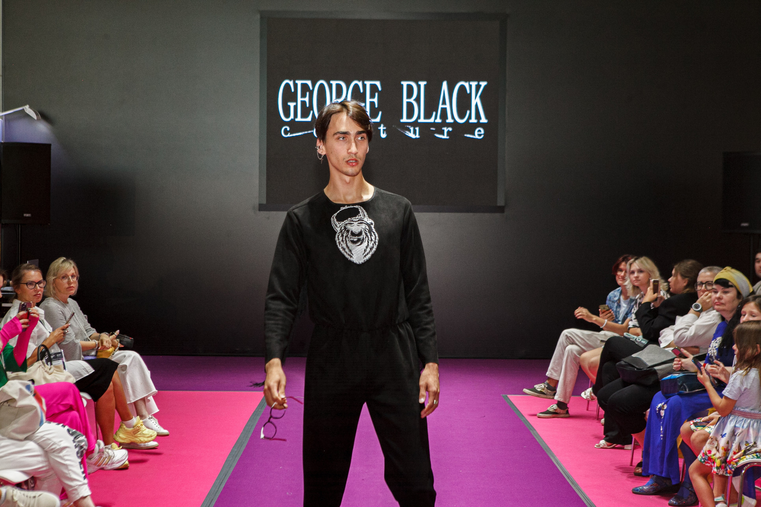  "Модели в ярких нарядах проходят по подиуму во время показа мод, освещенные яркими софитами и окруженные восторженными зрителями."