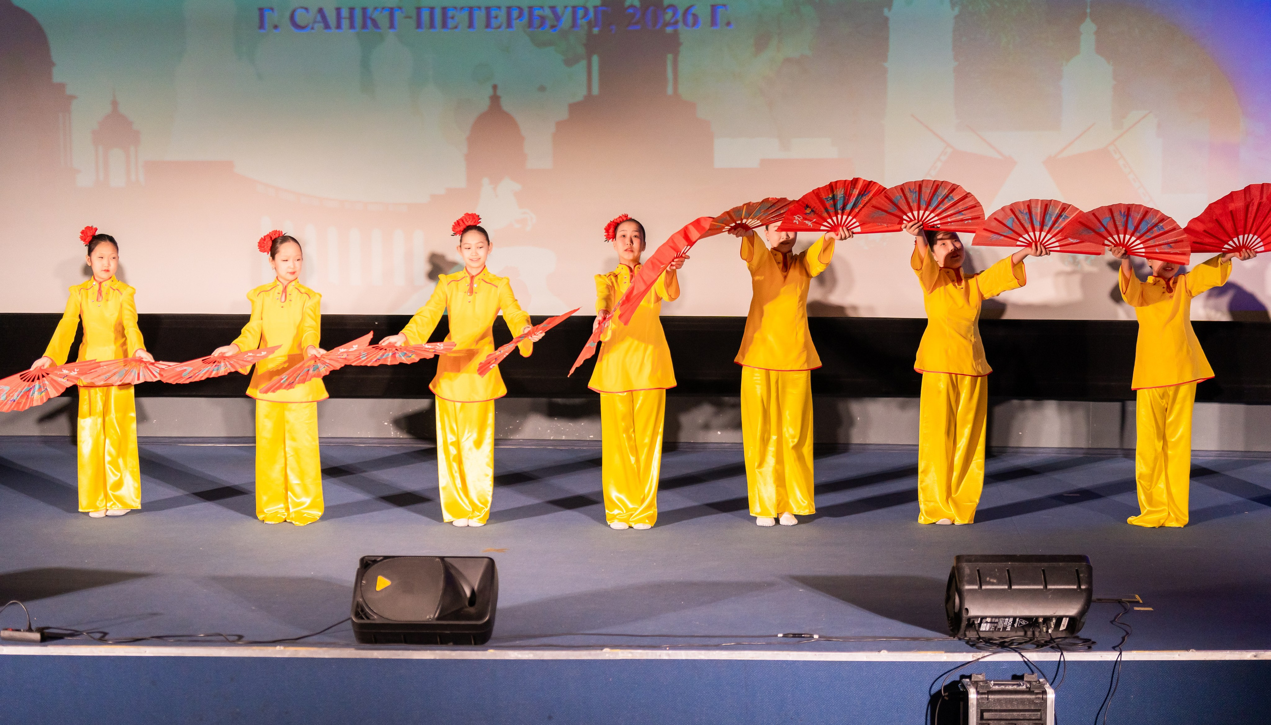 Девочки в жёлтых китайских костюмах с красными веерами на сцене XII Международного фольклорного фестиваля «Сохраняем традиции» Санкт-Петербург 2026. Фото: Мальцев Евгений, evrednix.ru.