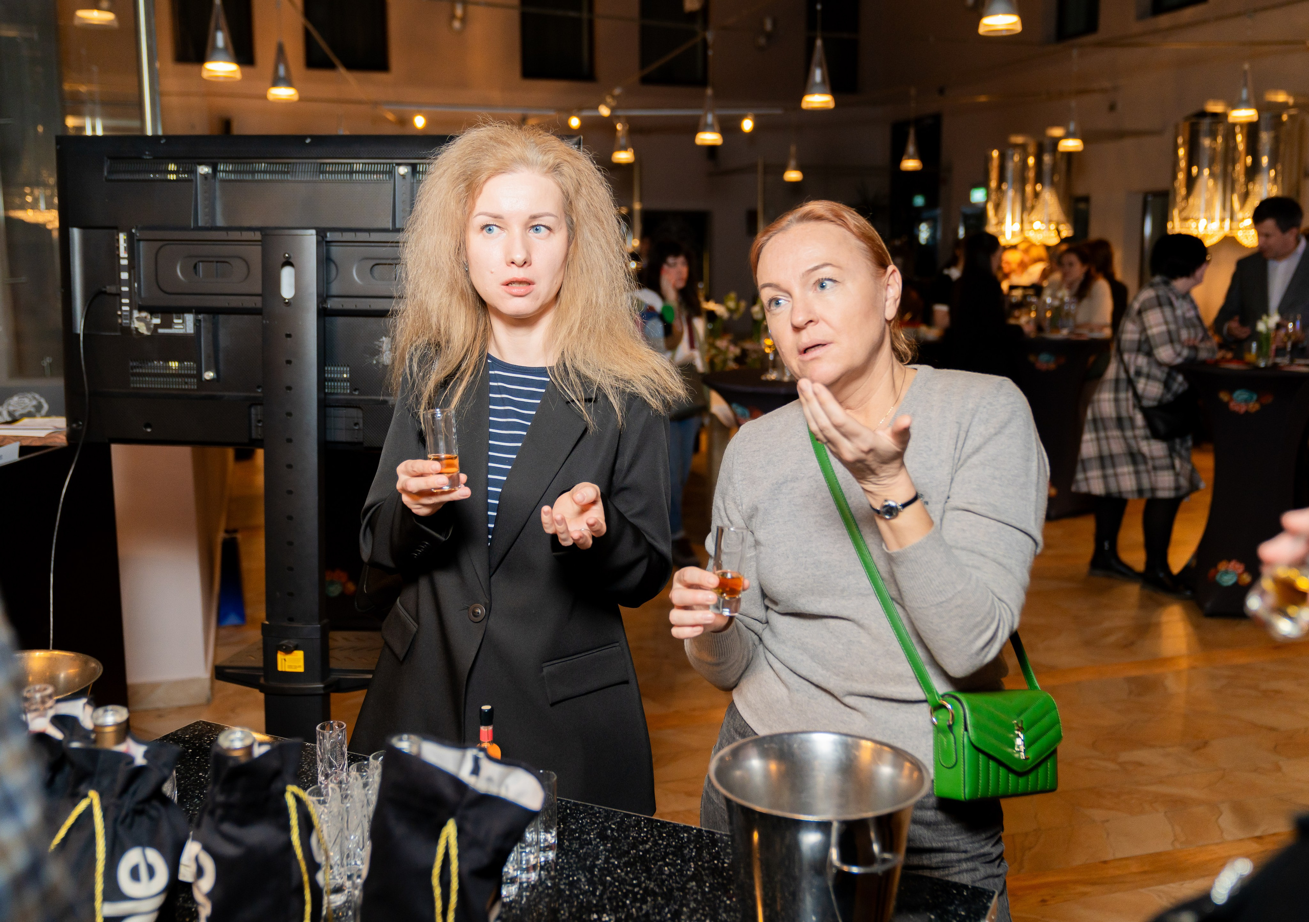 Две гостьи вечеринки в Radisson Sonya держат дегустационные бокалы и обсуждают напитки. Женщина в чёрном блейзере и женщина в сером свитере. Фотограф Евгений Мальцев, альбом Radisson Sonya, evrednix.ru.
